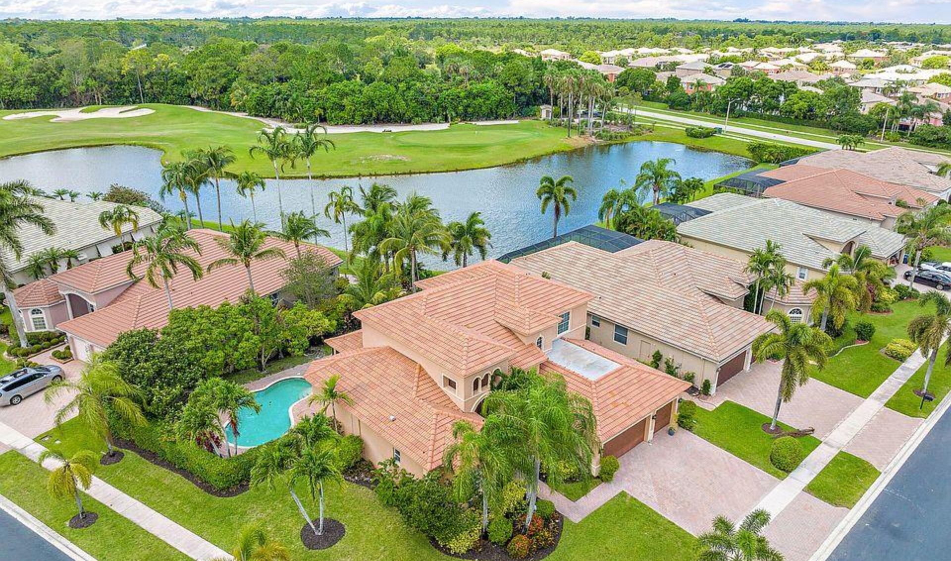 Aerial view of tile-roofed homes beside a lake and golf course, with palms and a backyard pool.