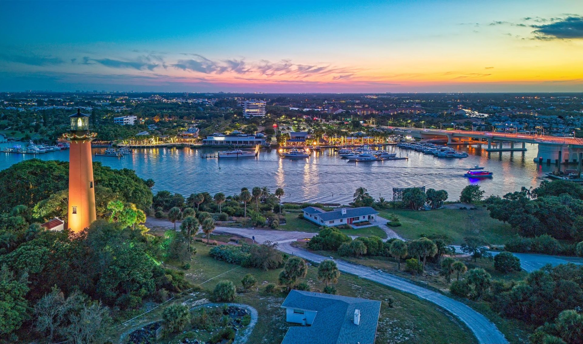 Jupiter Inlet waterfront at dusk, with lighthouse, marina, boats, and bridge.