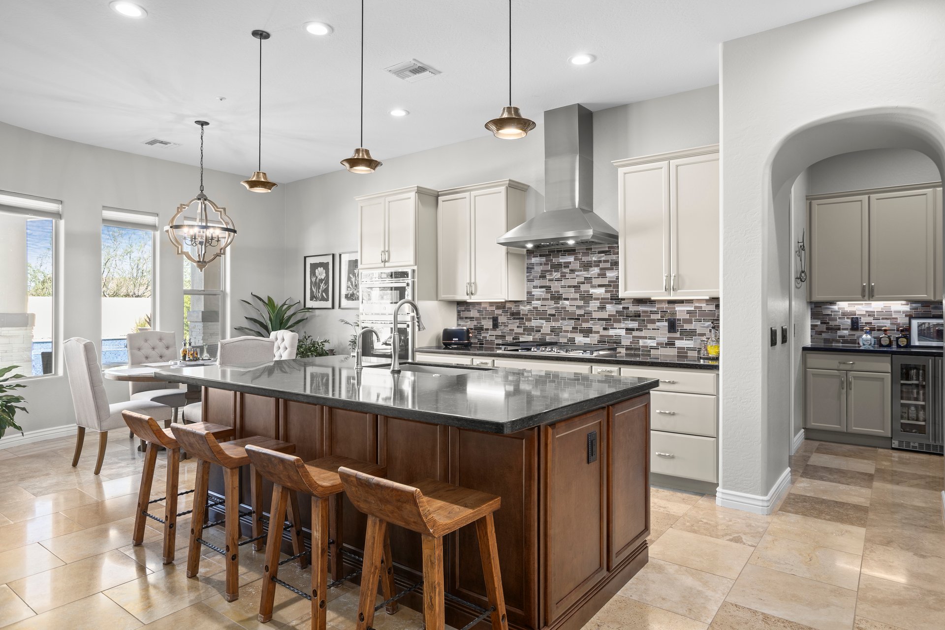 Bright open kitchen with a large island, bar stools, pendant lights, white cabinetry, tiled backsplash, stainless hood, and adjoining dining nook.