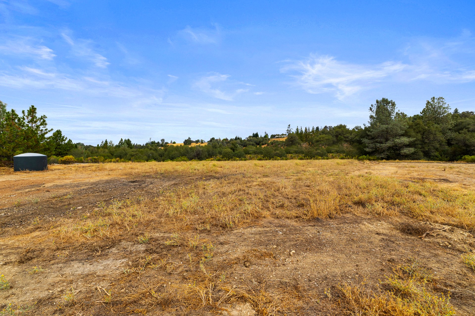 Open dry lot bordered by trees, with a dark water tank at the left edge.