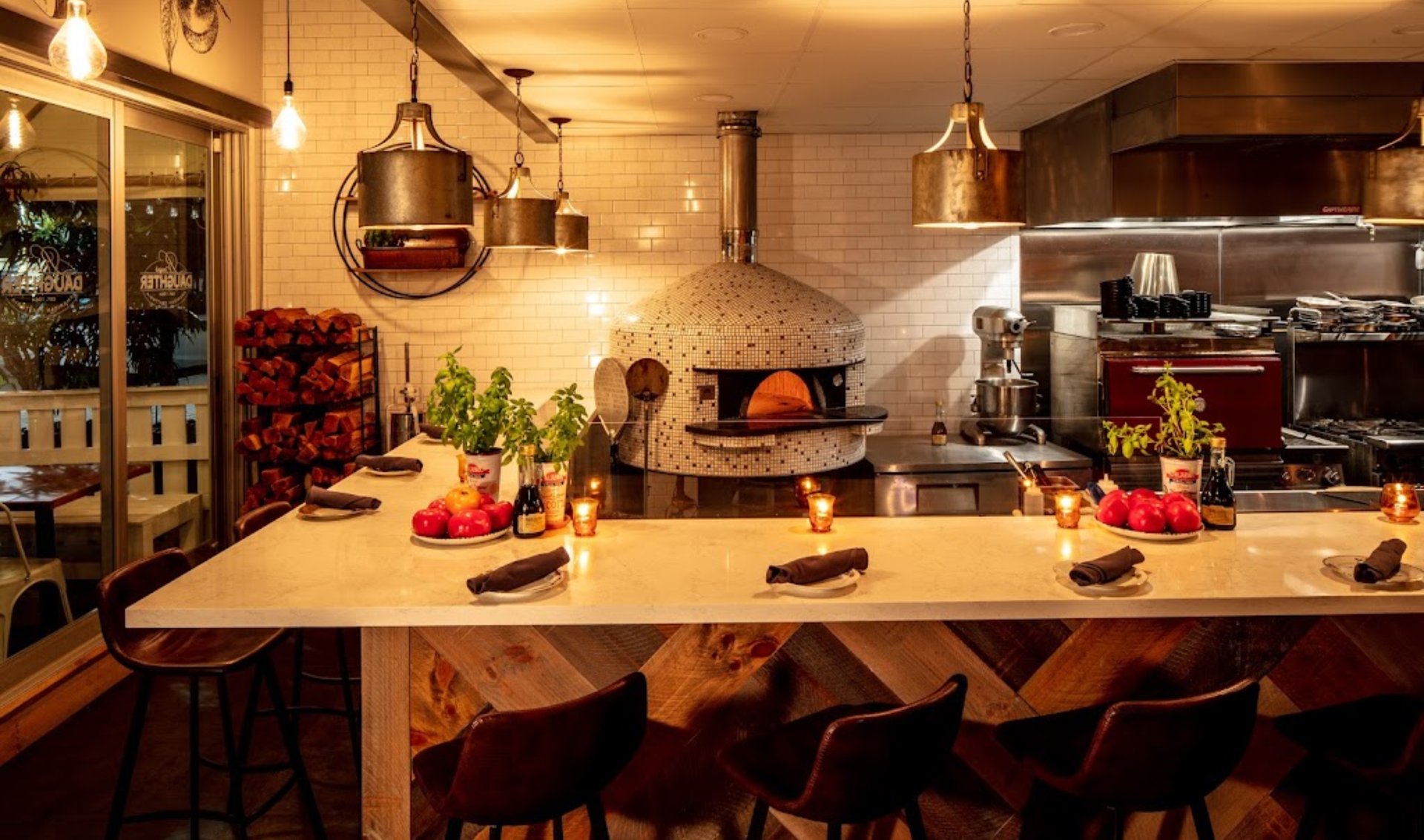 Restaurant counter and stools facing a tiled wood-fired oven and open kitchen.