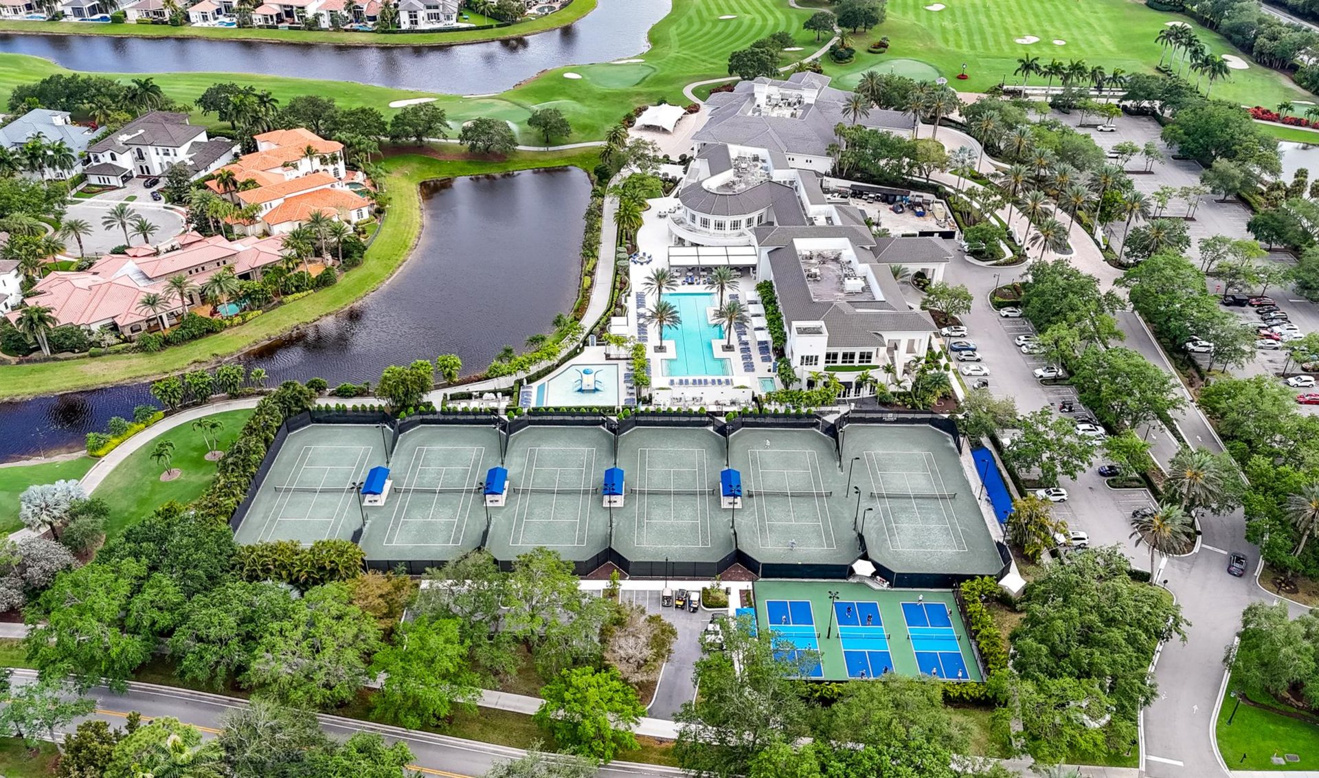 Aerial view of a country club with clubhouse, pools, courts, golf course, and lakeside homes.