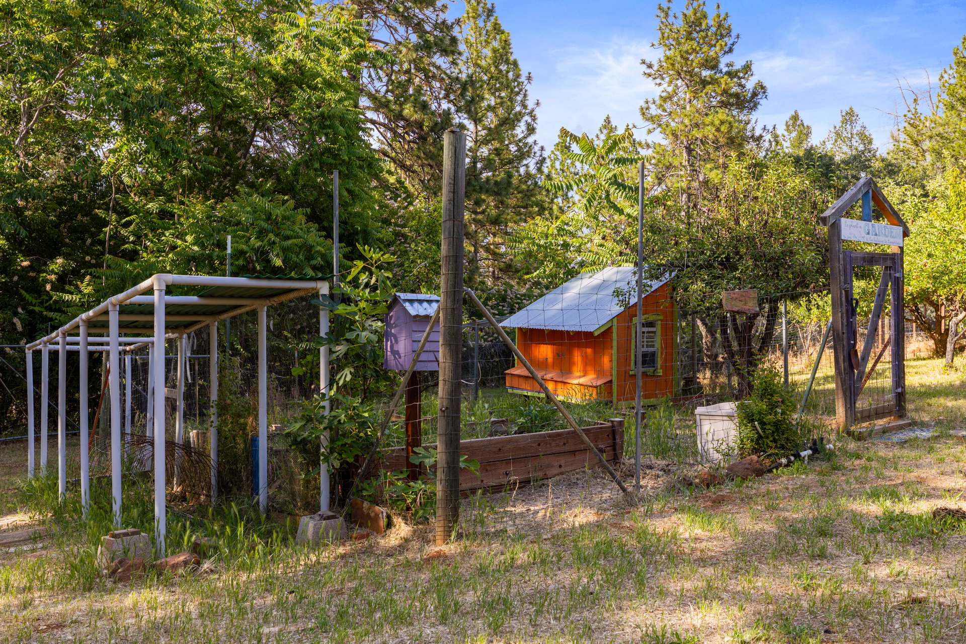 Fenced garden with raised beds, a small orange outbuilding, and a covered structure beneath mature trees.