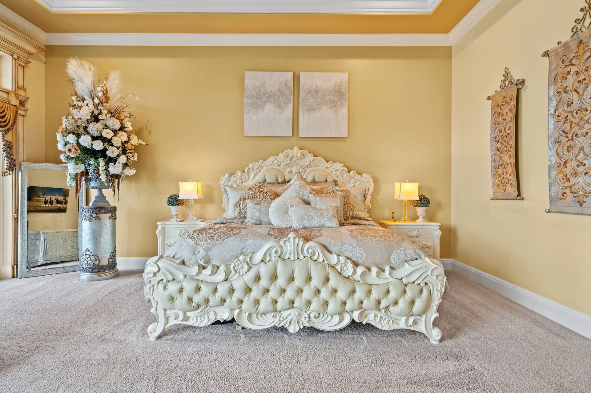 Bedroom with an ornate white bed, gold walls, and a tall floral arrangement beside a floor mirror.