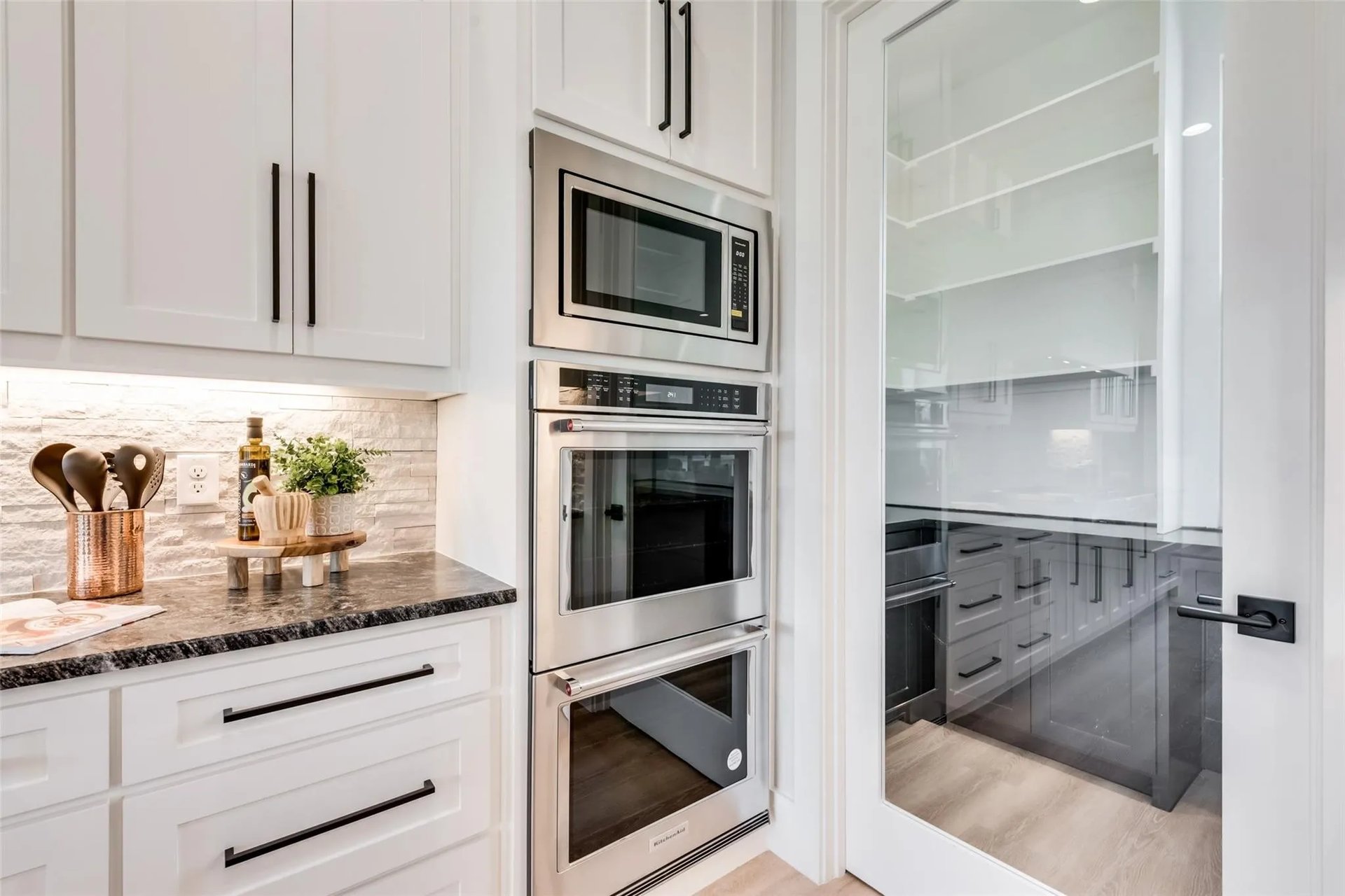 Kitchen with white cabinetry, stainless-steel double ovens, dark countertops, and a glass-door pantry.