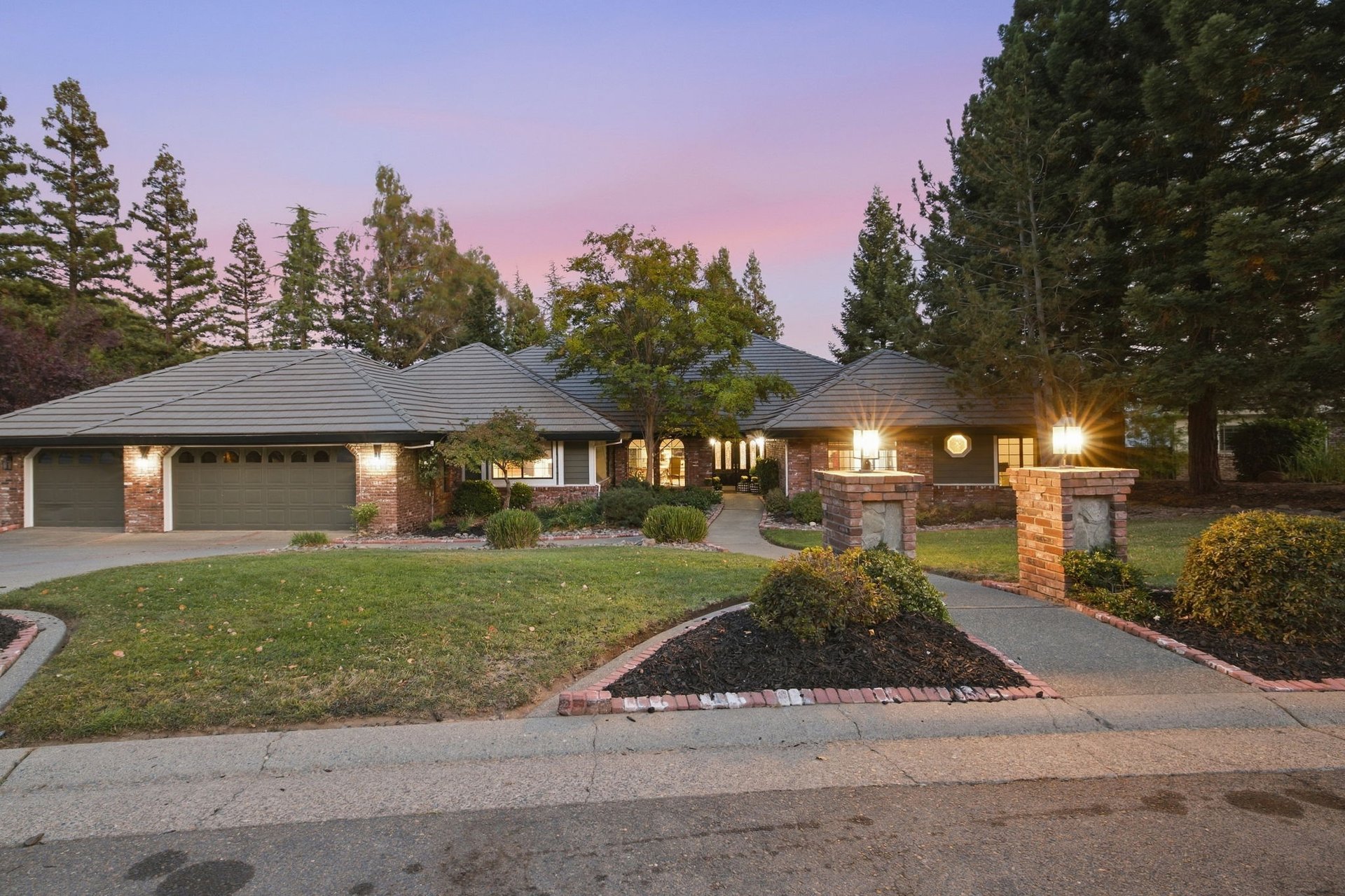 Single-story brick home with a gray tile roof, landscaped yard, and illuminated entry at dusk.