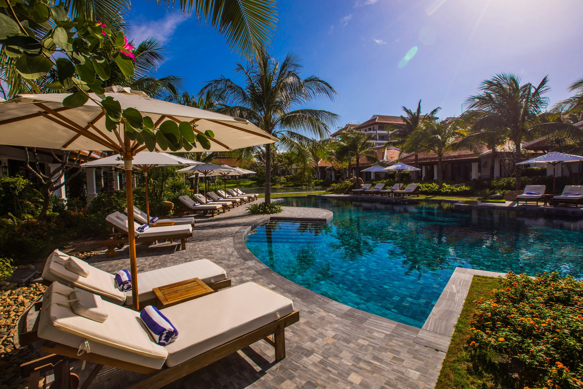 Luxurious poolside setting with lounge chairs, umbrellas, palm trees, and resort-style villas in the background.