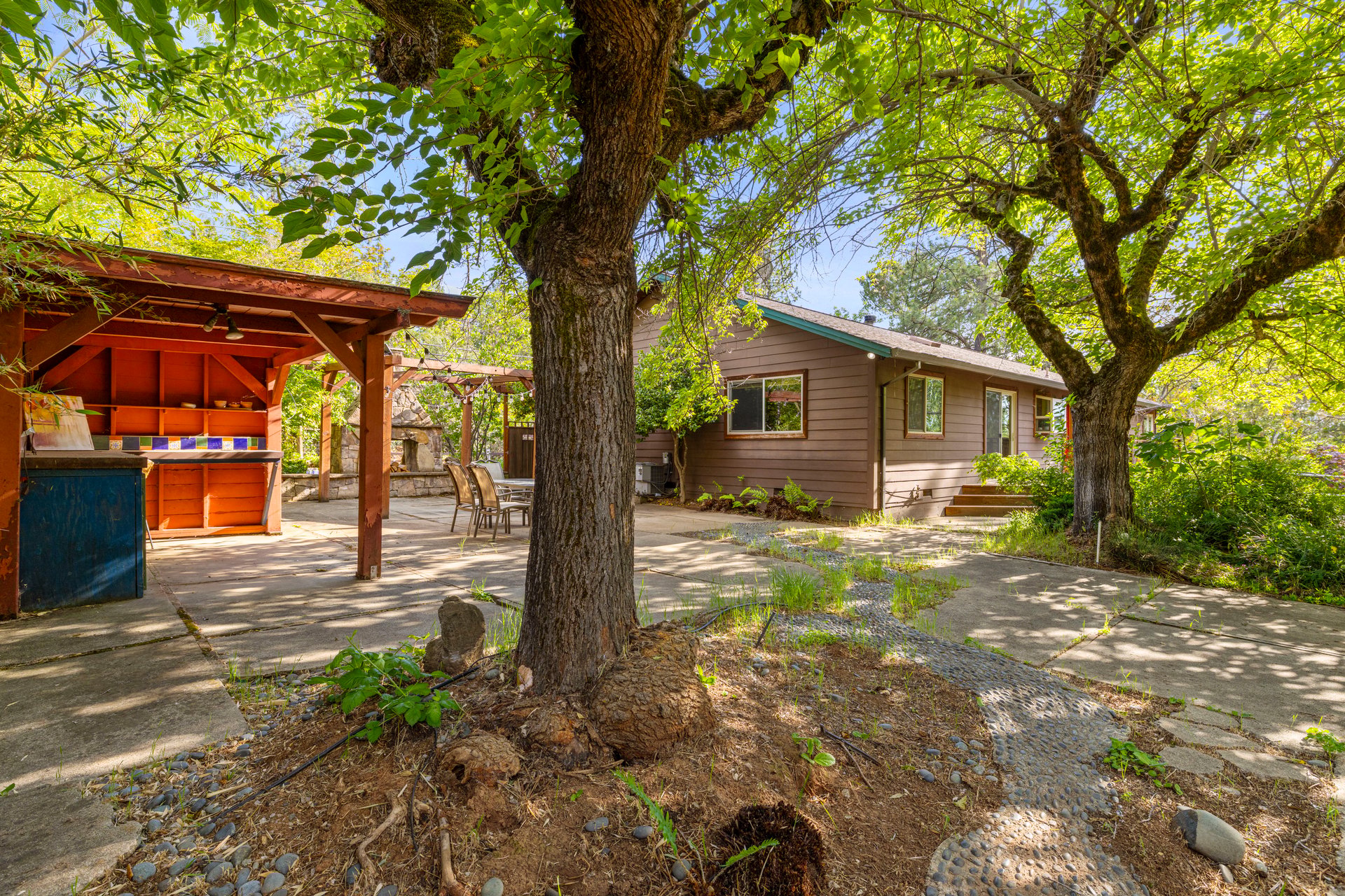 Single-story brown house beside a covered outdoor kitchen and patio shaded by mature trees.