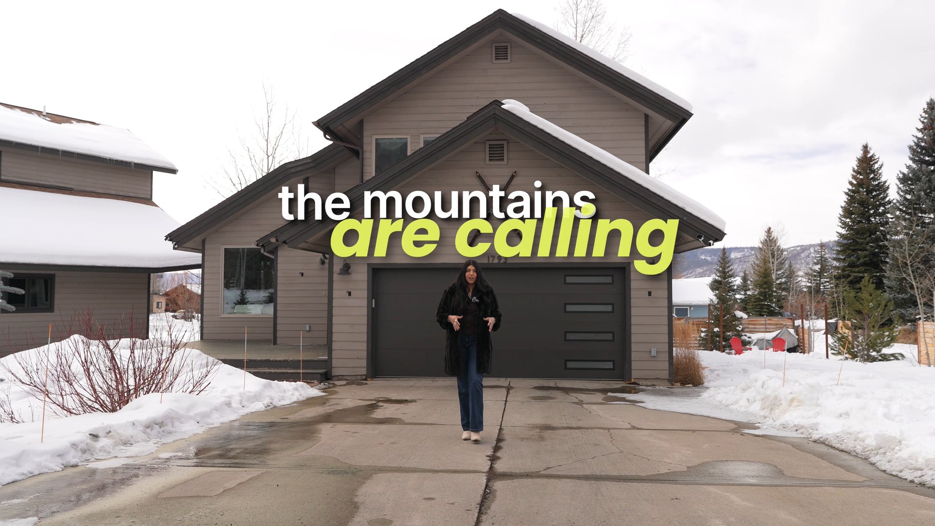 Two-story mountain home with a dark double garage, snow-covered yard, and a woman in the driveway.