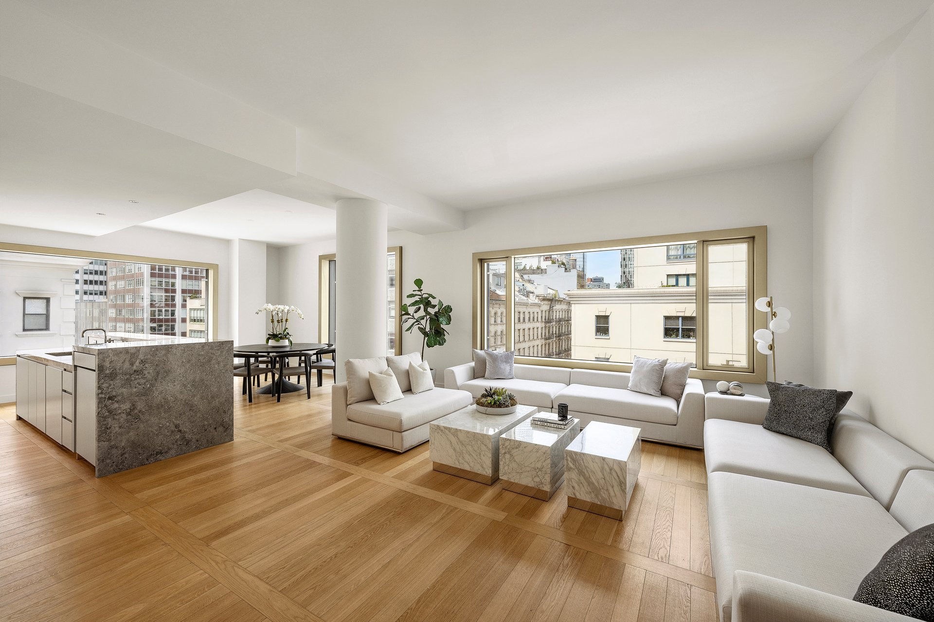 Open-plan living room with white sectional sofas, marble tables, and large windows overlooking city buildings.