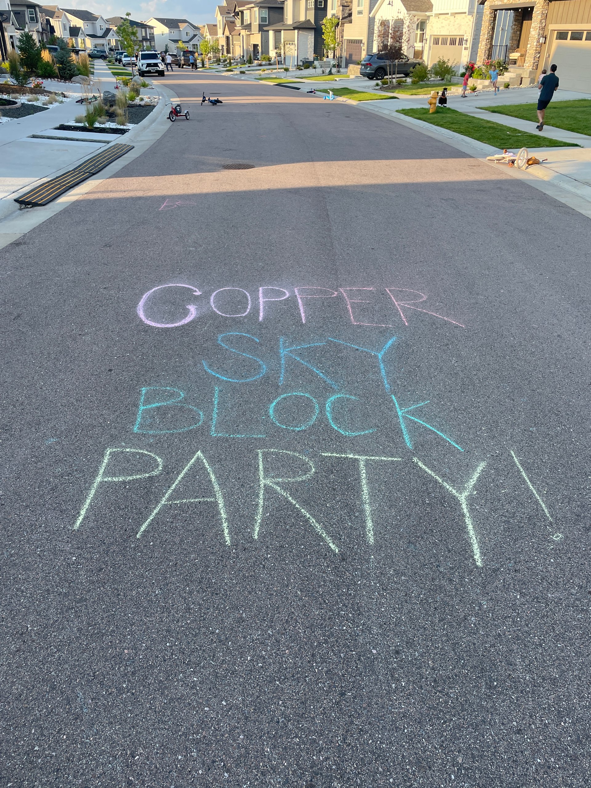 Residential street with modern homes, people outdoors, and colorful block party chalk lettering on the asphalt.