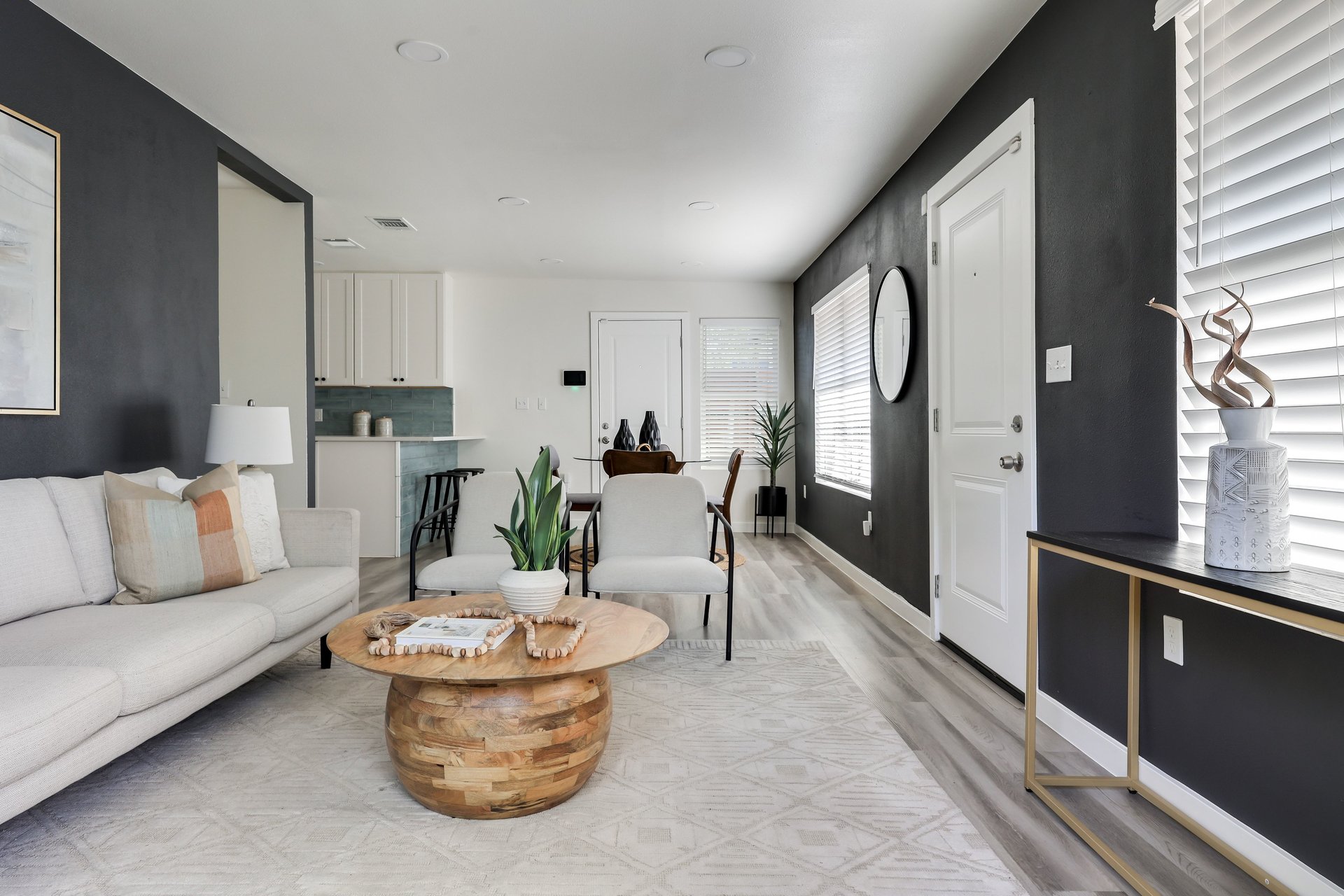Staged living room with a white sofa, armchairs, round wood coffee table, and open kitchen.