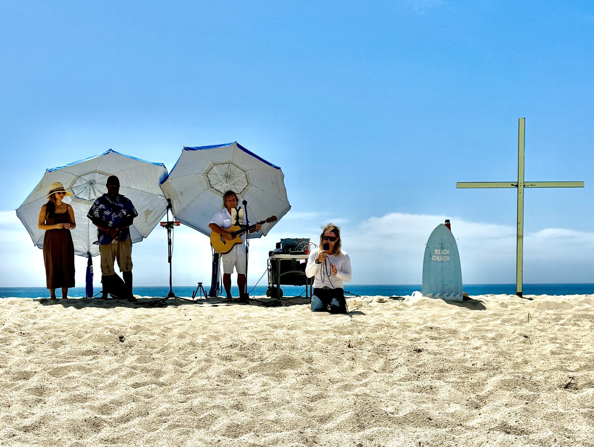 Musicians and a singer at a beach worship gathering beside a cross and Beach Church sign.