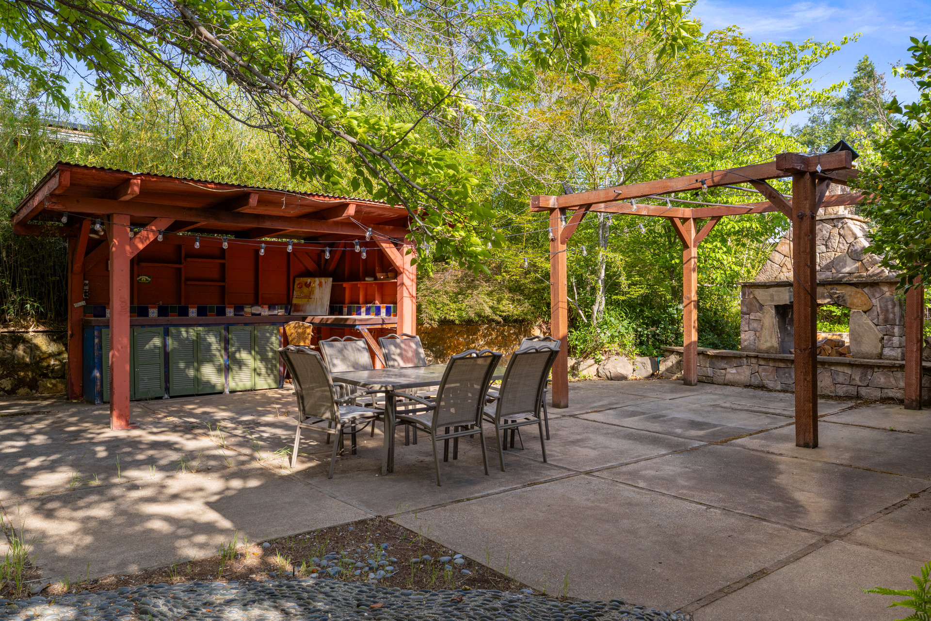Outdoor patio with a pergola, dining set, covered kitchen, and stone fireplace among leafy trees.