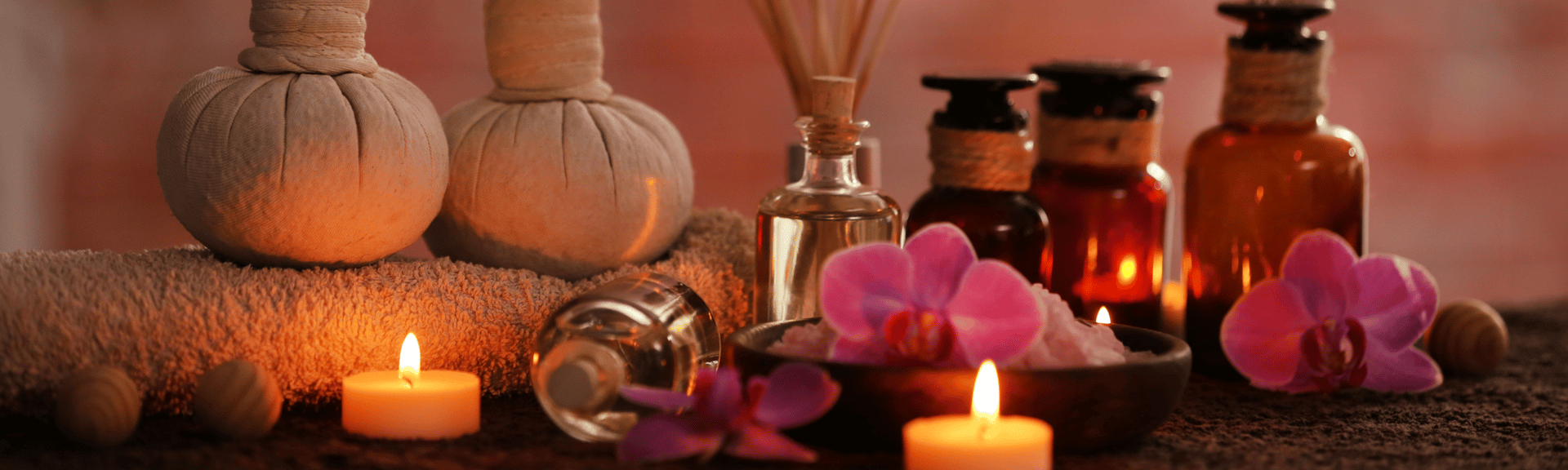 A serene spa setting featuring herbal pouches, essential oil bottles, candles, and pink orchids on a textured surface.