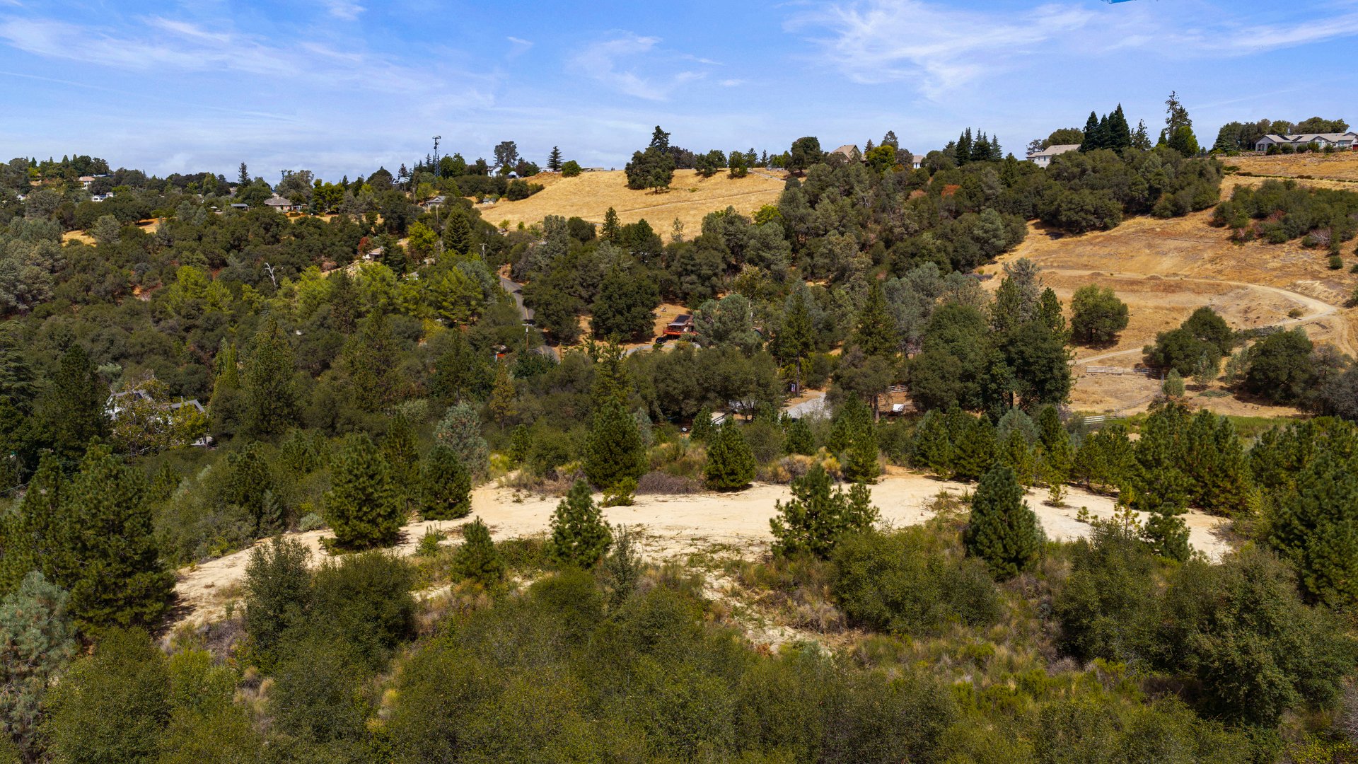 Aerial view of a wooded hillside property with a broad cleared area and homes beyond.