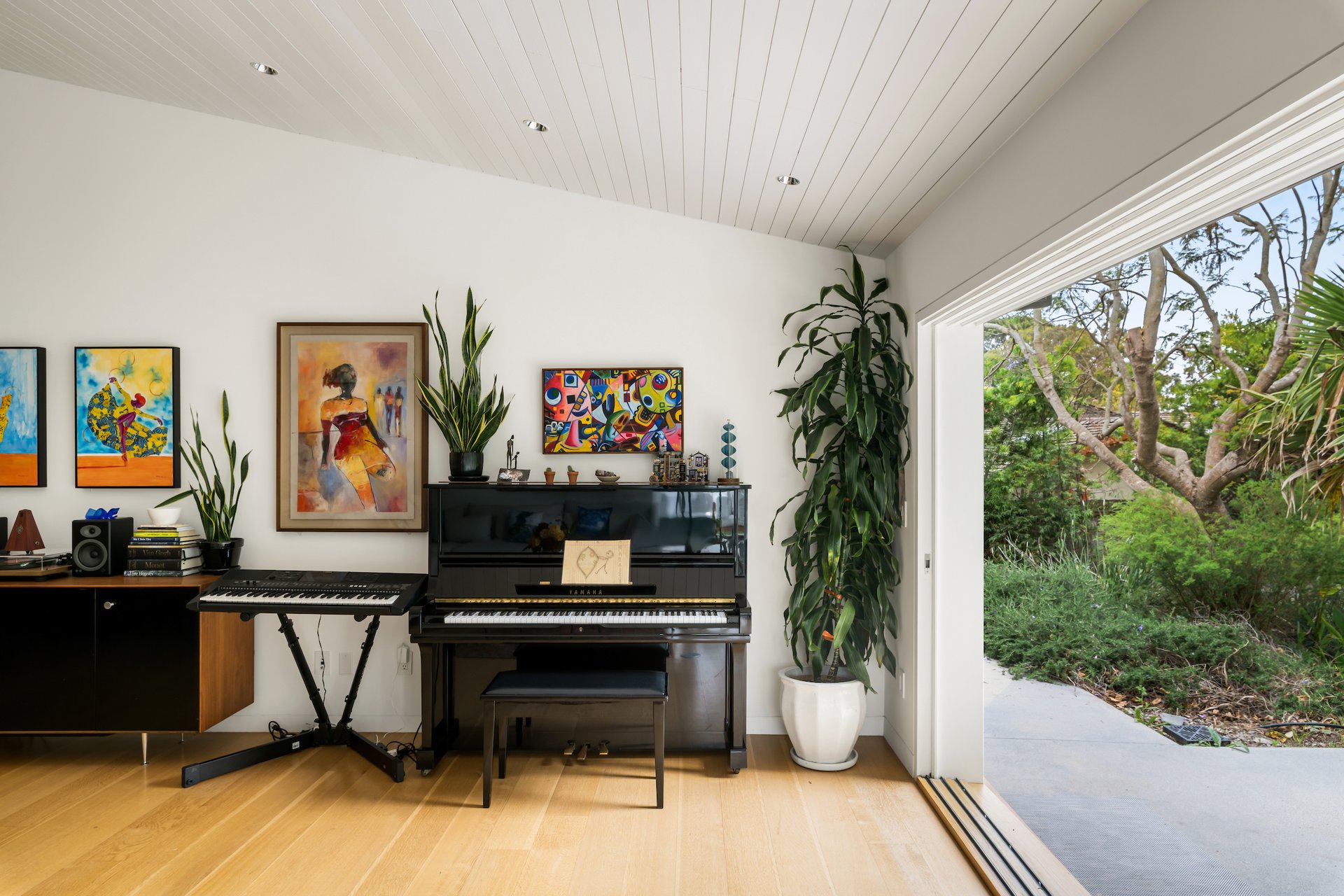Music room with a black piano, keyboard, colorful artwork, and an open sliding door to the garden.