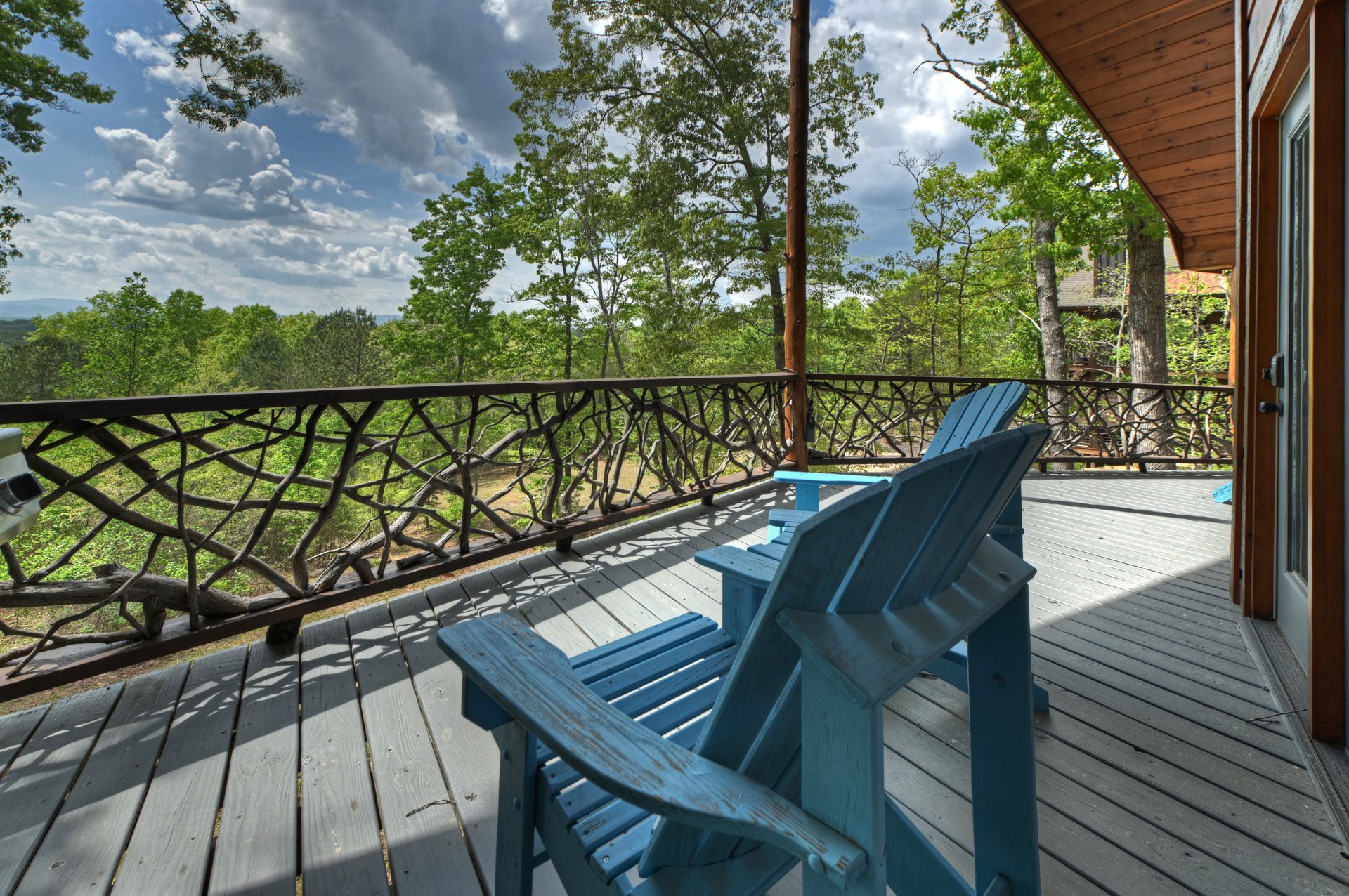 Covered mountain-home deck with blue Adirondack chairs, rustic branch railing, and distant ridges.