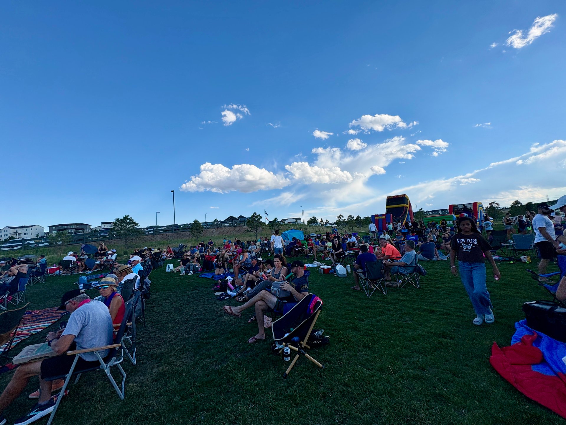 Neighbors gather on a grassy field with lawn chairs and inflatable attractions.