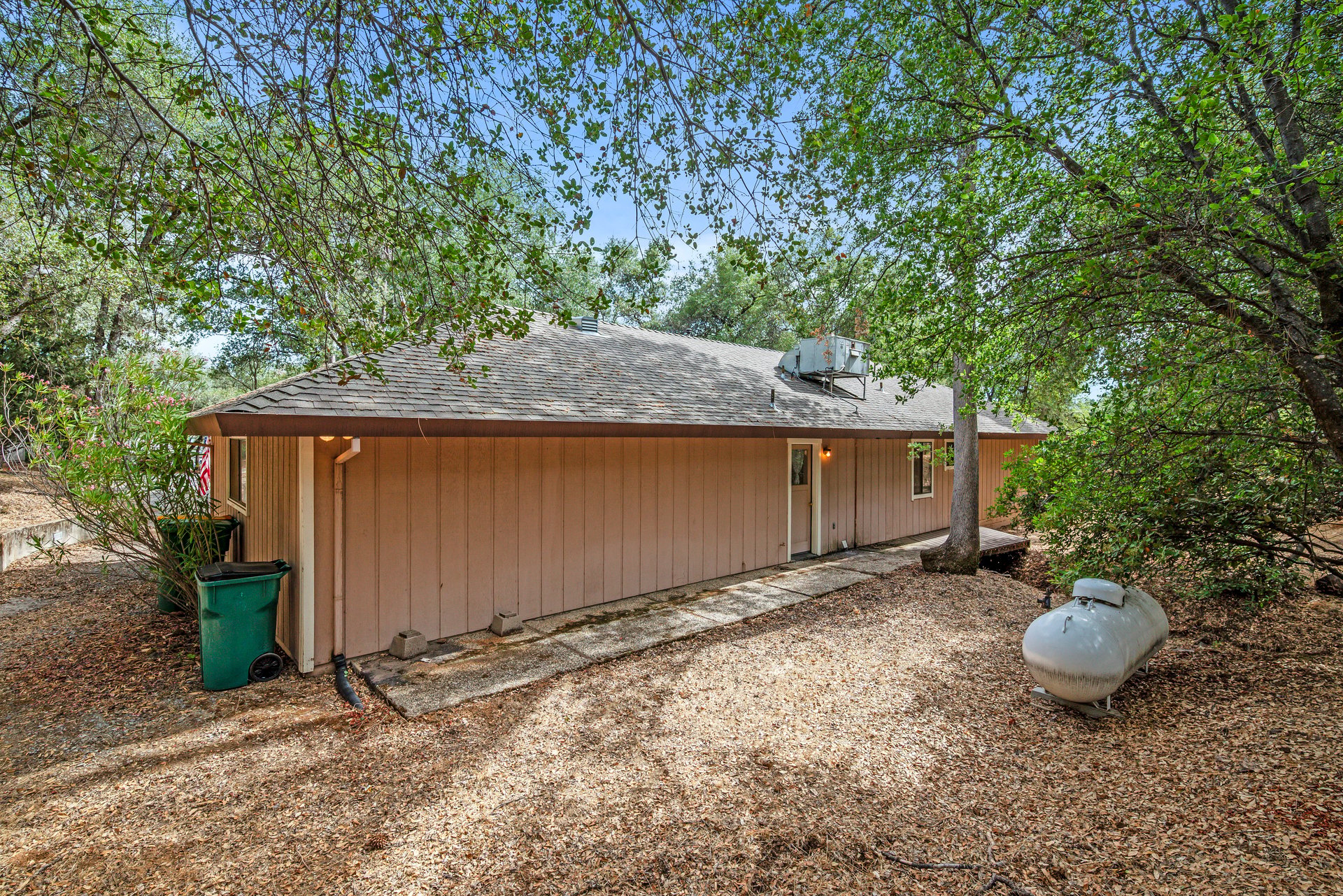 Single-story tan-sided home surrounded by trees, with a concrete walkway and propane tank.