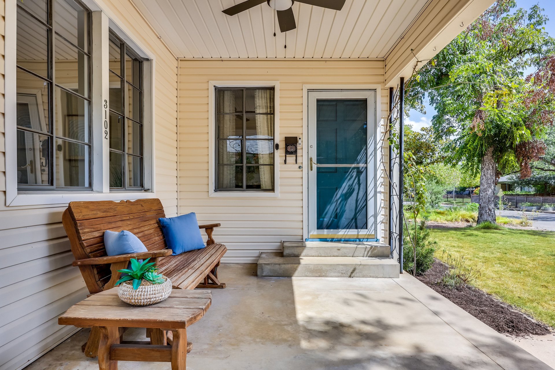 Covered front porch with teal door, wooden bench, and leafy yard.
