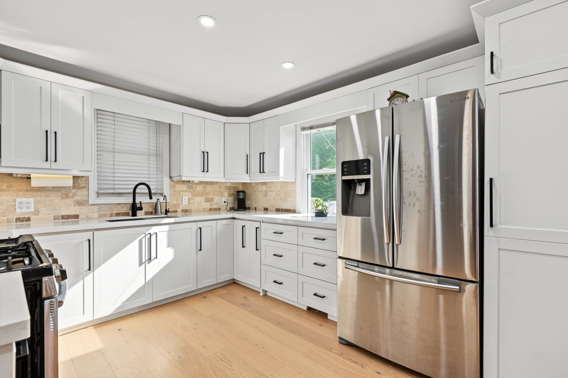 Bright kitchen with white cabinetry, a stainless steel refrigerator, stone tile backsplash, and light wood floors.
