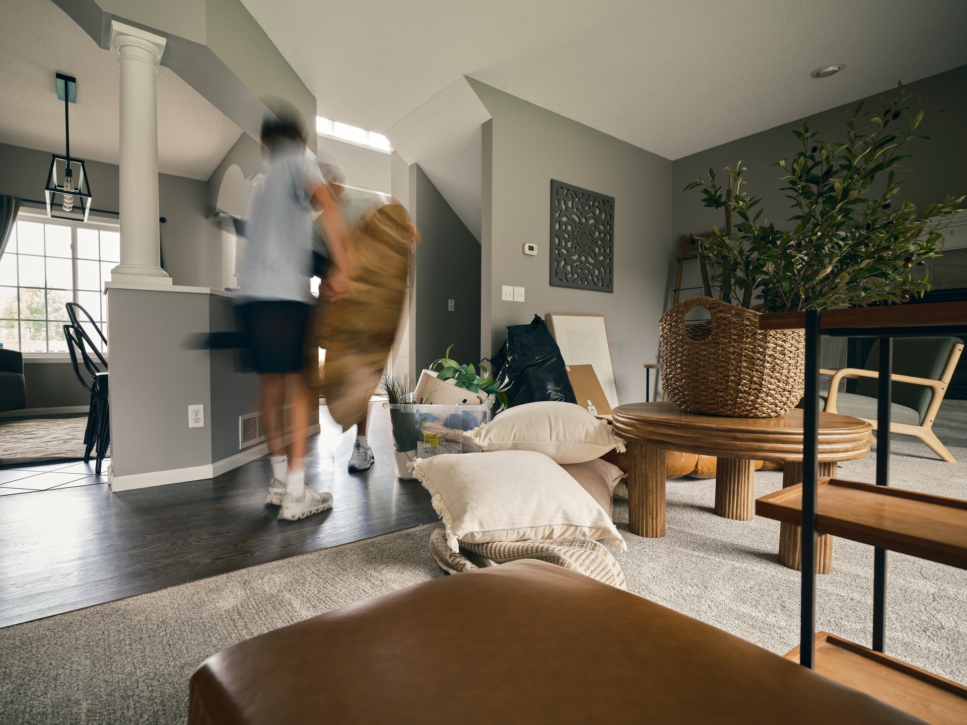 Open-plan living room being staged, with people arranging cushions beside a round wood table and tall plant.