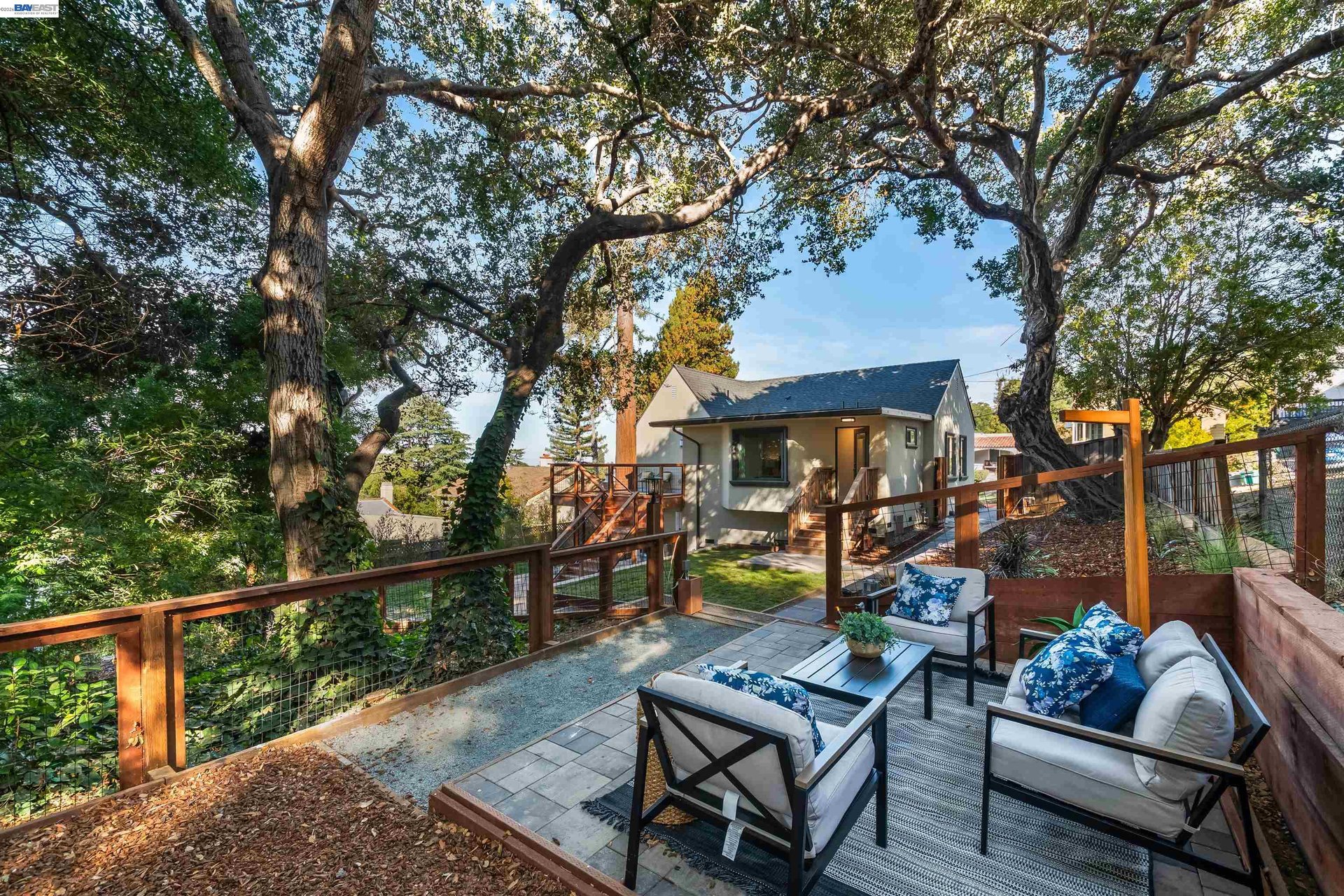 Furnished stone patio and rear of a house beneath large mature trees.
