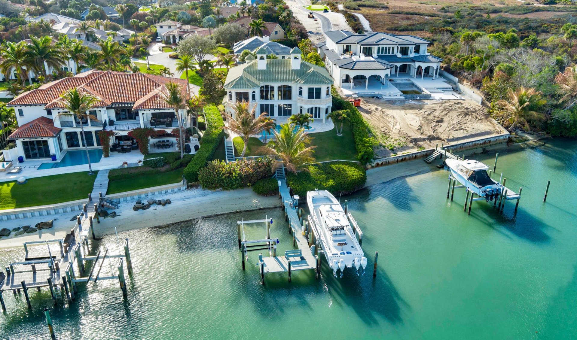 Aerial view of waterfront homes and private docks along a turquoise canal, with boats and a neighboring construction site.