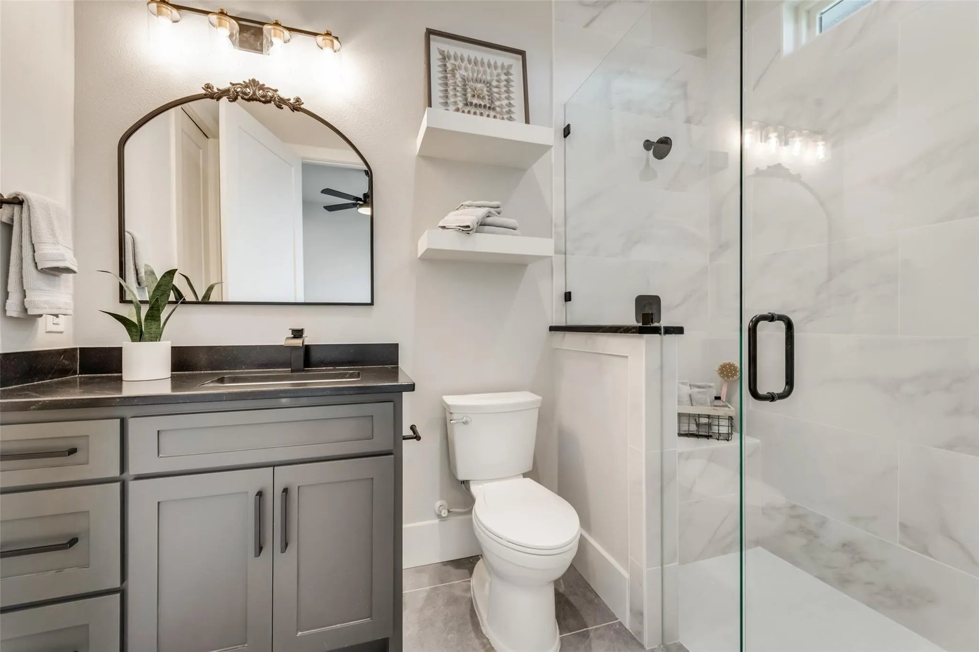 Bathroom with a gray vanity, arched mirror, and glass-enclosed marble-look shower.