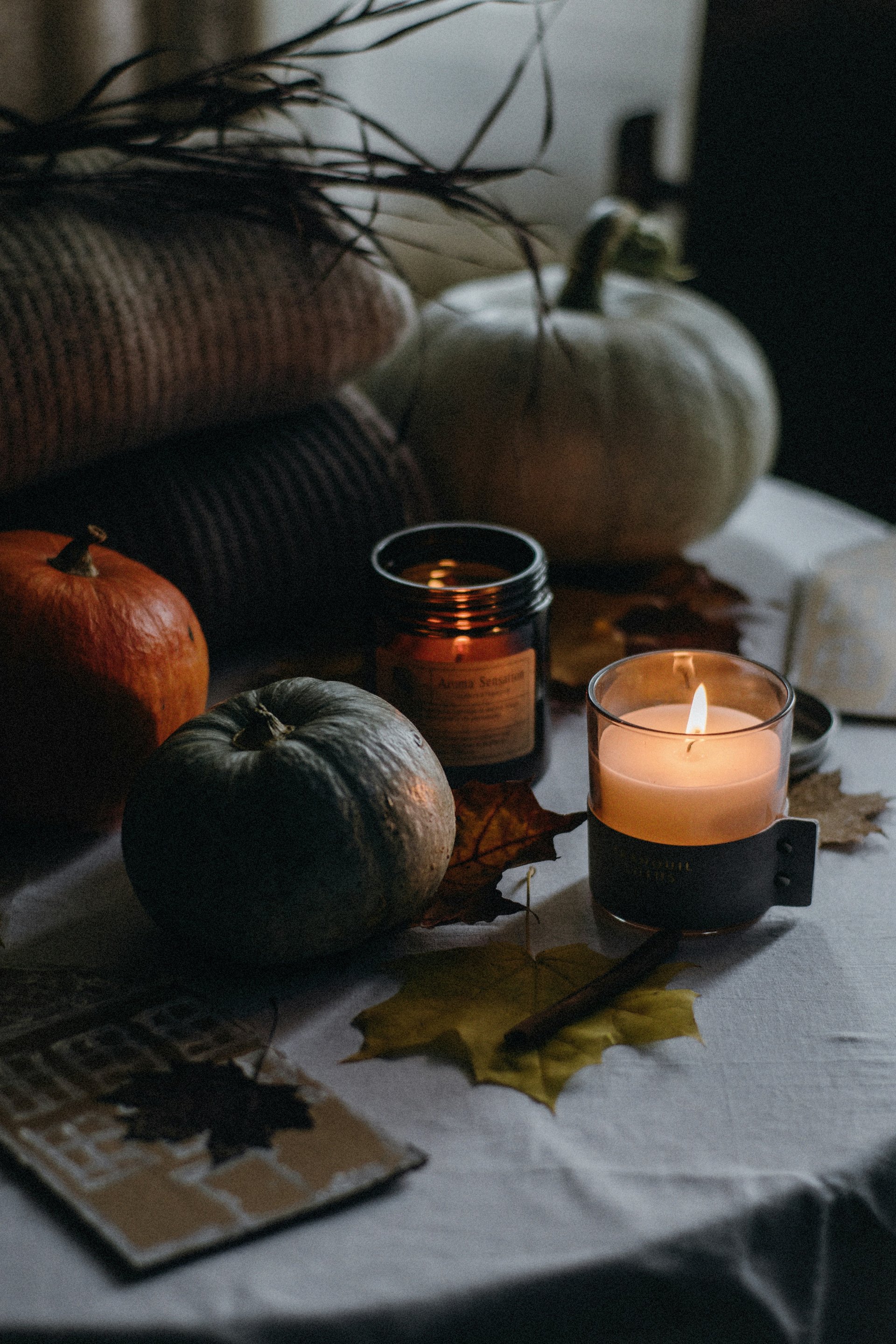 Lit candle and pumpkins with fallen autumn leaves on a table.
