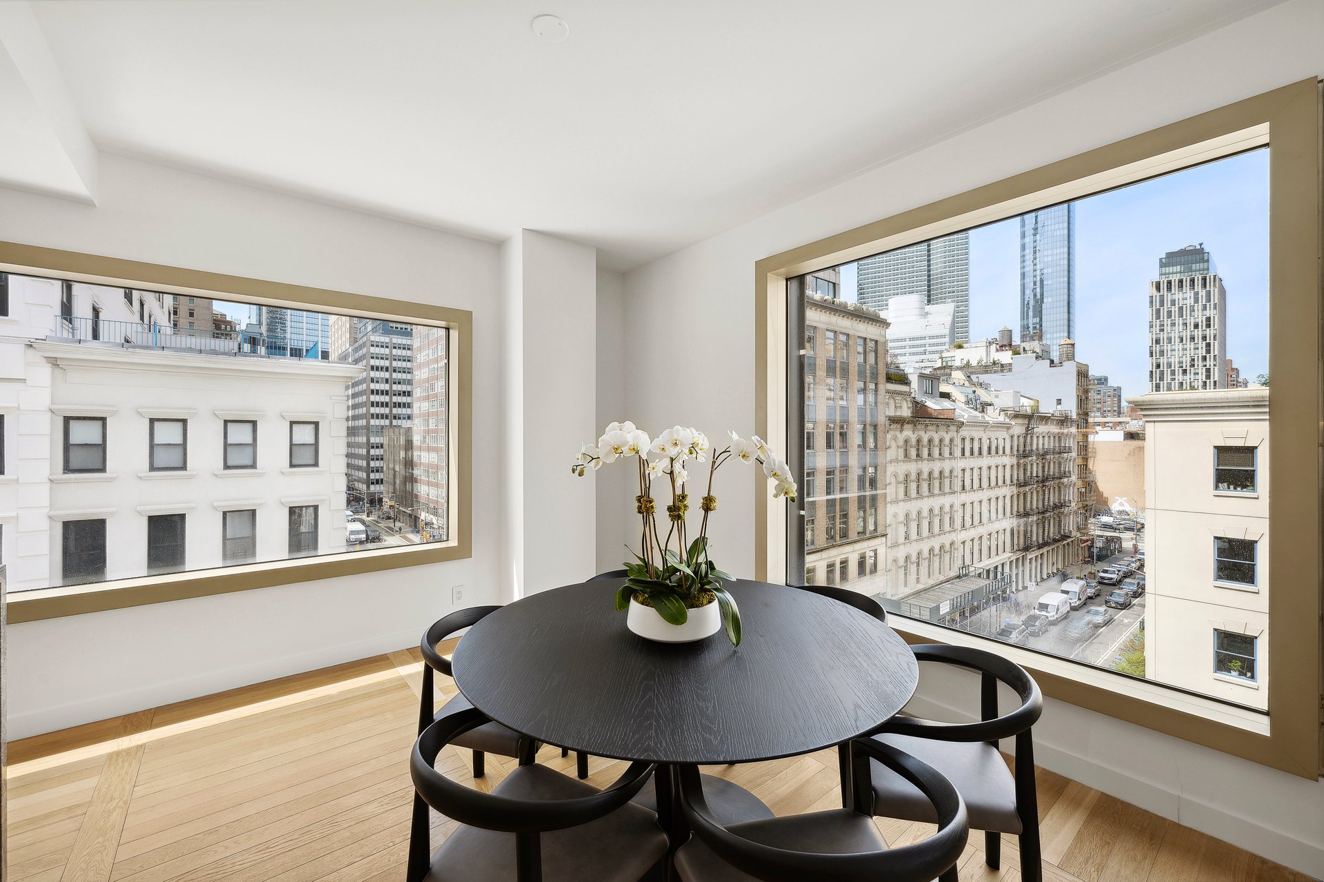 Dining area with a round table and orchids beside large windows overlooking city buildings.