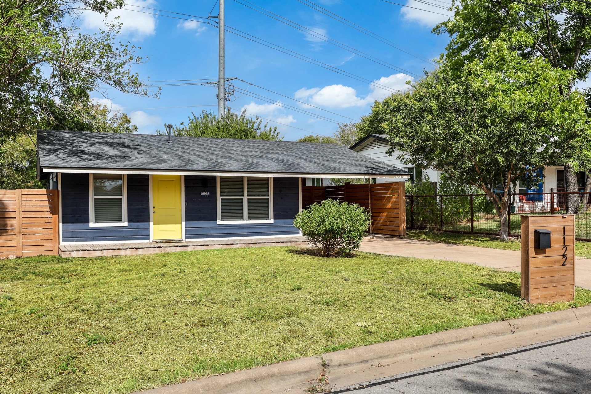 Single-story blue house with a bright yellow front door, lawn, and wooden fencing.