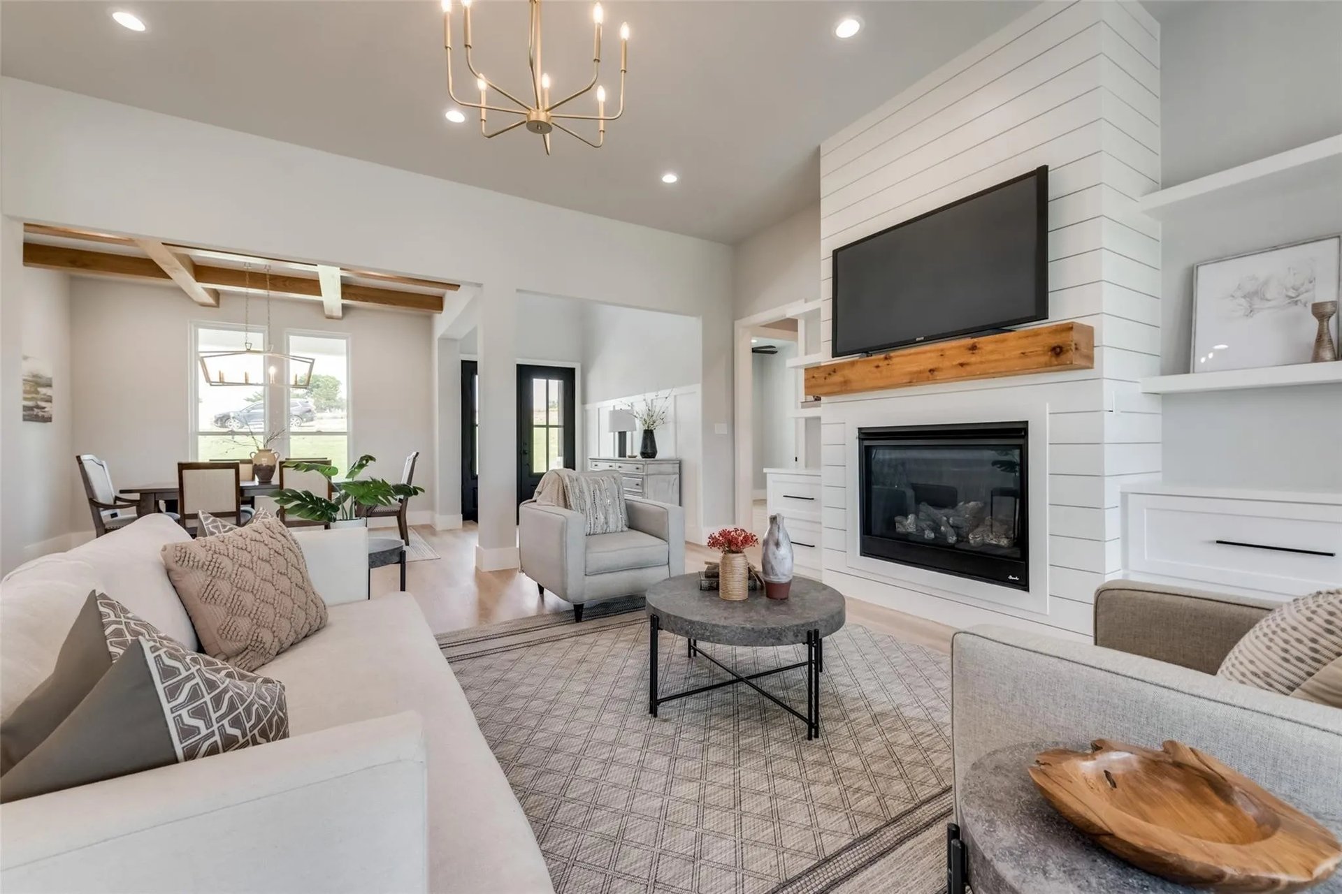 Staged living room with a shiplap fireplace, mounted TV, exposed beams, and adjoining dining area.