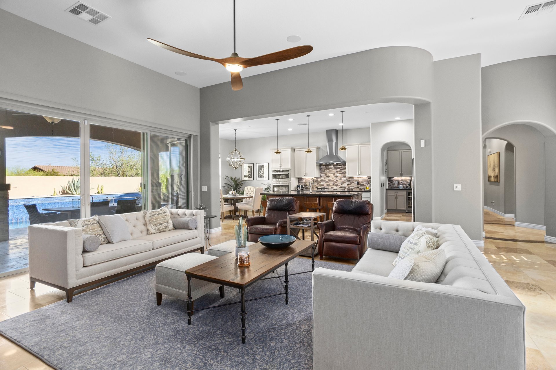 Bright staged living room with two sofas, recliners, coffee tables, and a view into the kitchen and out to a patio and pool through large sliding doors.
