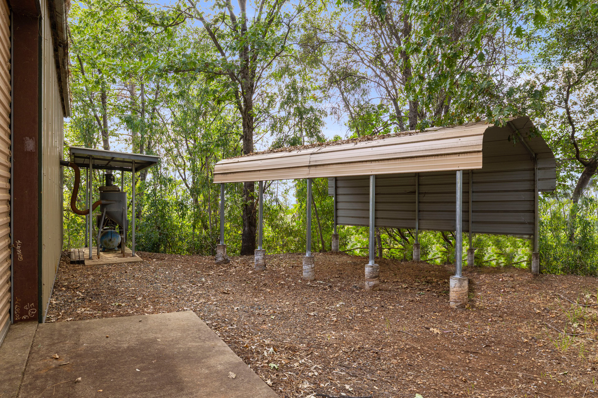 Raised metal-roofed structure on concrete piers in a wooded yard beside a building.