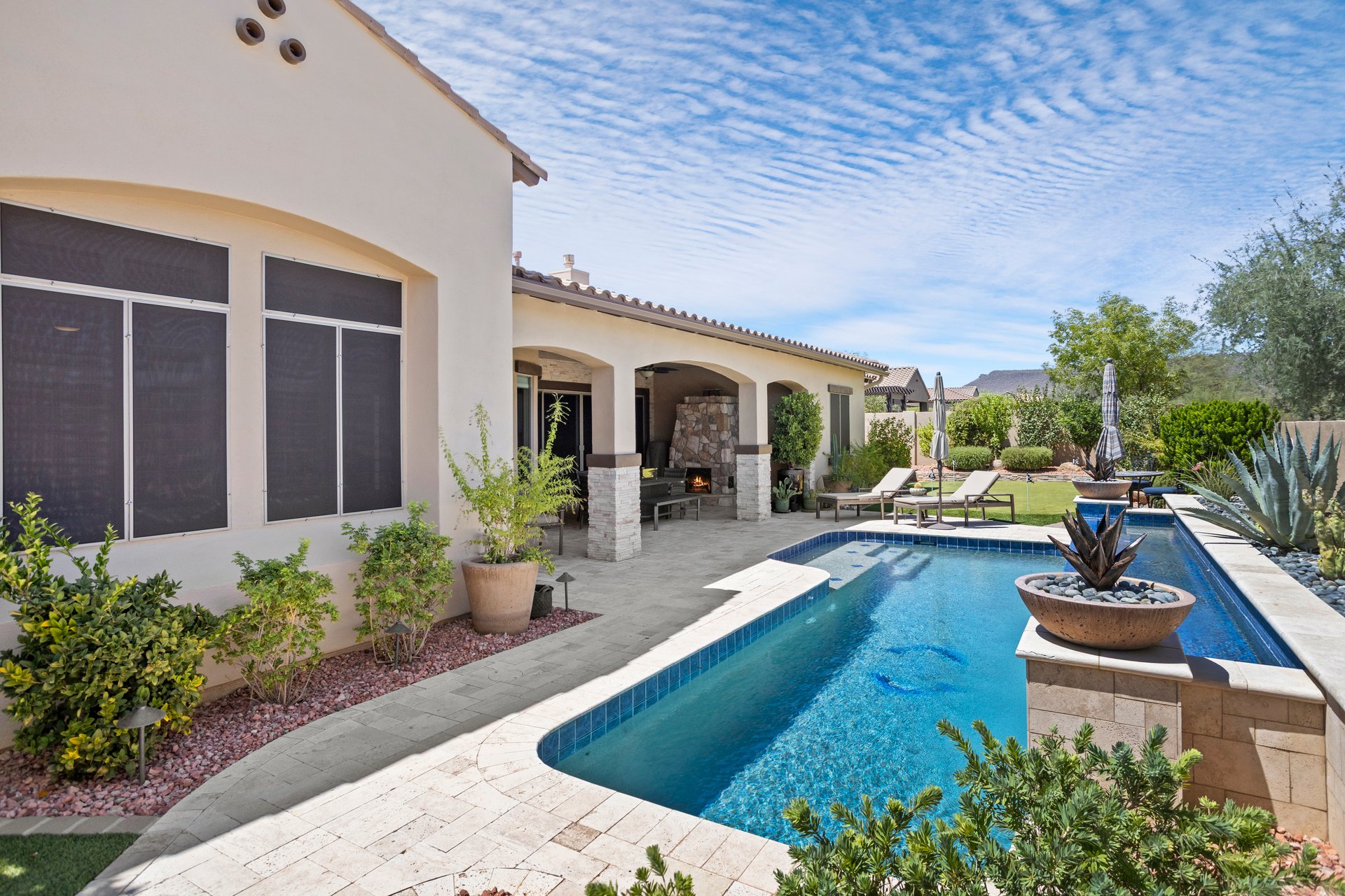 A landscaped backyard patio with a blue swimming pool, lounge chairs, covered outdoor fireplace, and desert-style plants beside a stucco house.