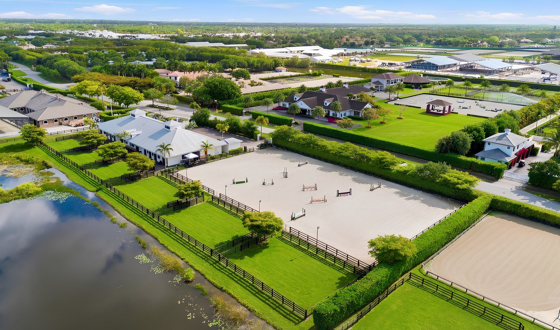 Aerial view of an equestrian property with a jump arena, barns, fenced paddocks, and a pond.