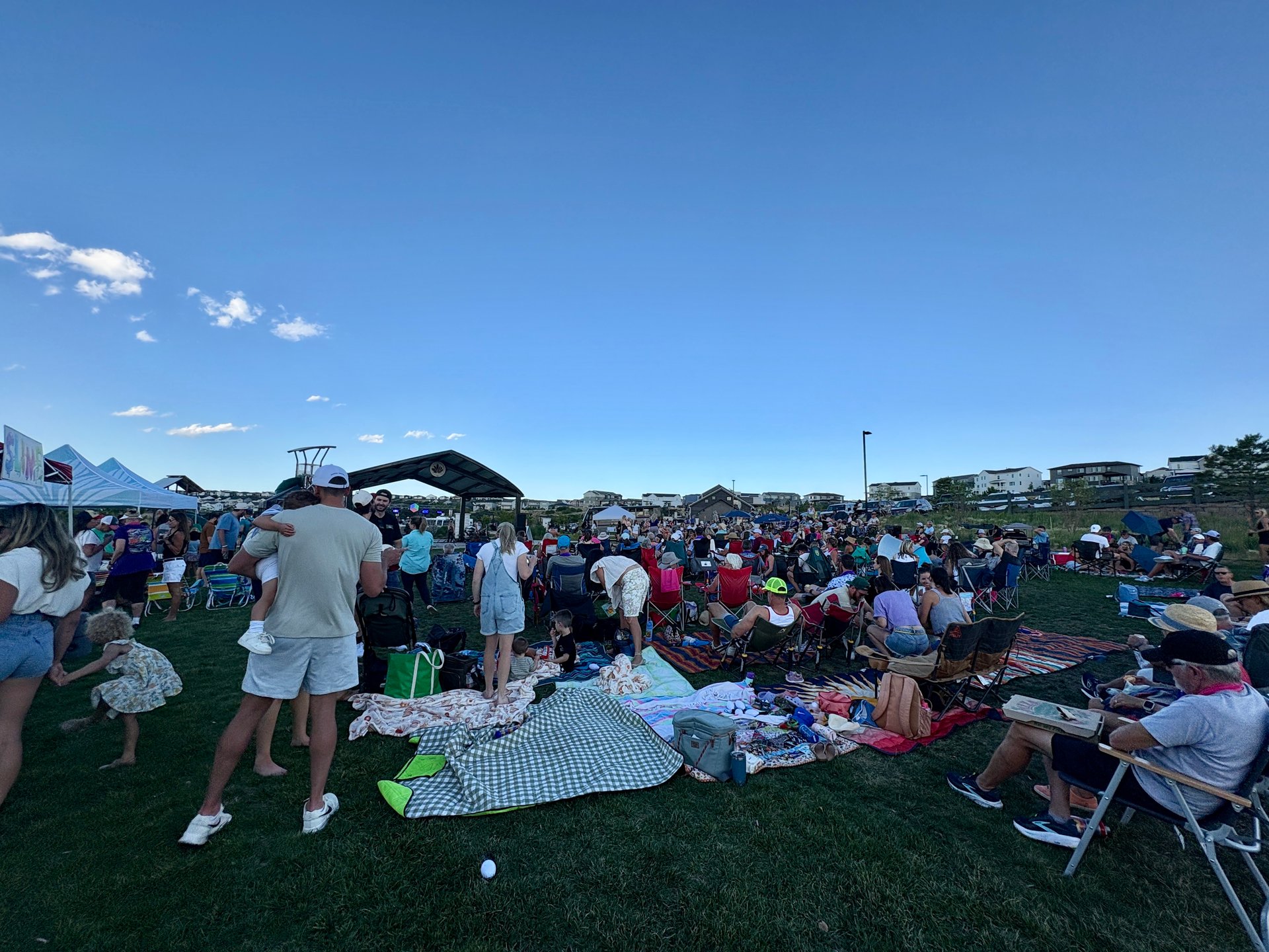 Neighbors gather on a grassy field for an outdoor community event, seated on blankets and folding chairs.