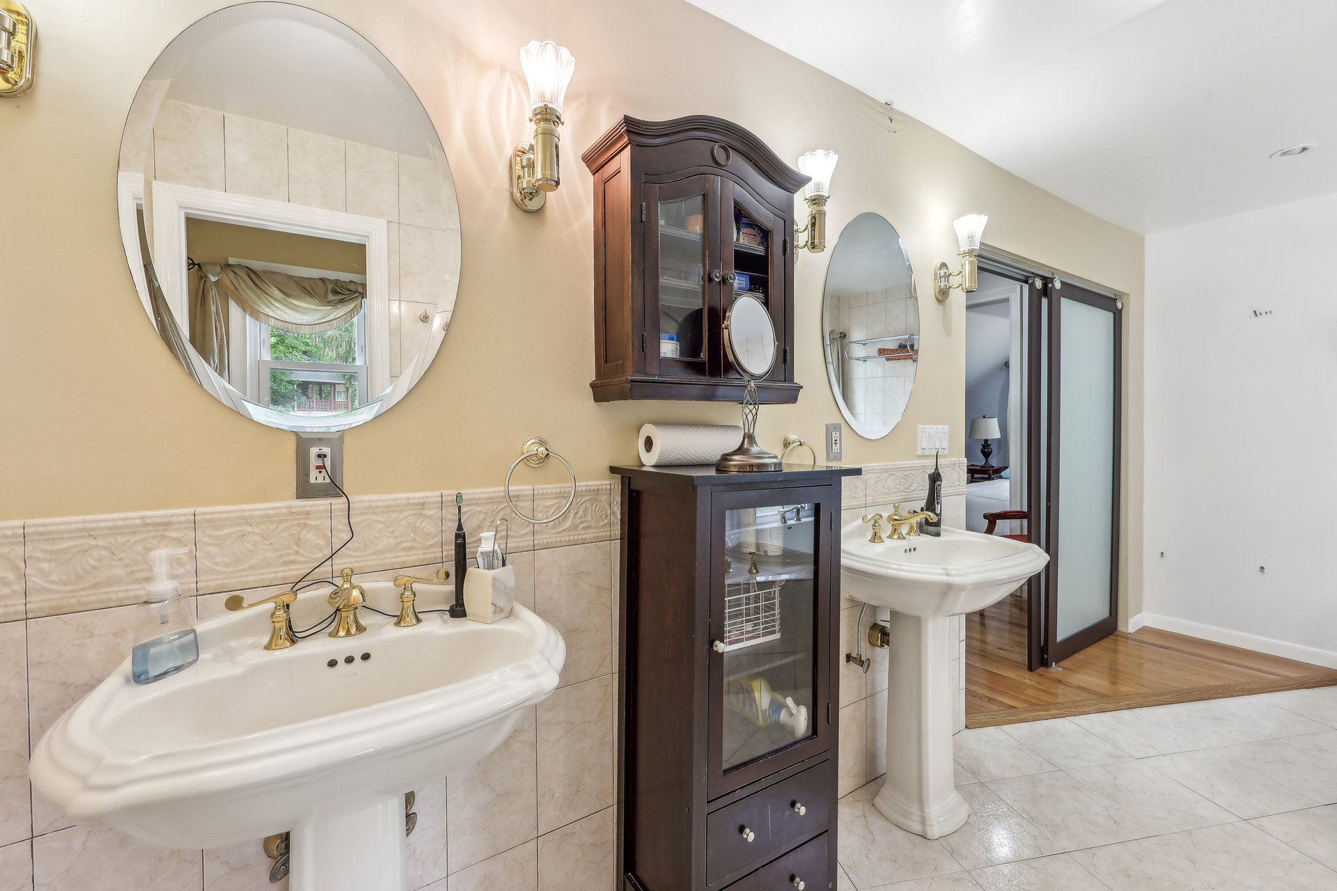 Bathroom with two pedestal sinks, oval mirrors, beige tile, and dark wood cabinets.