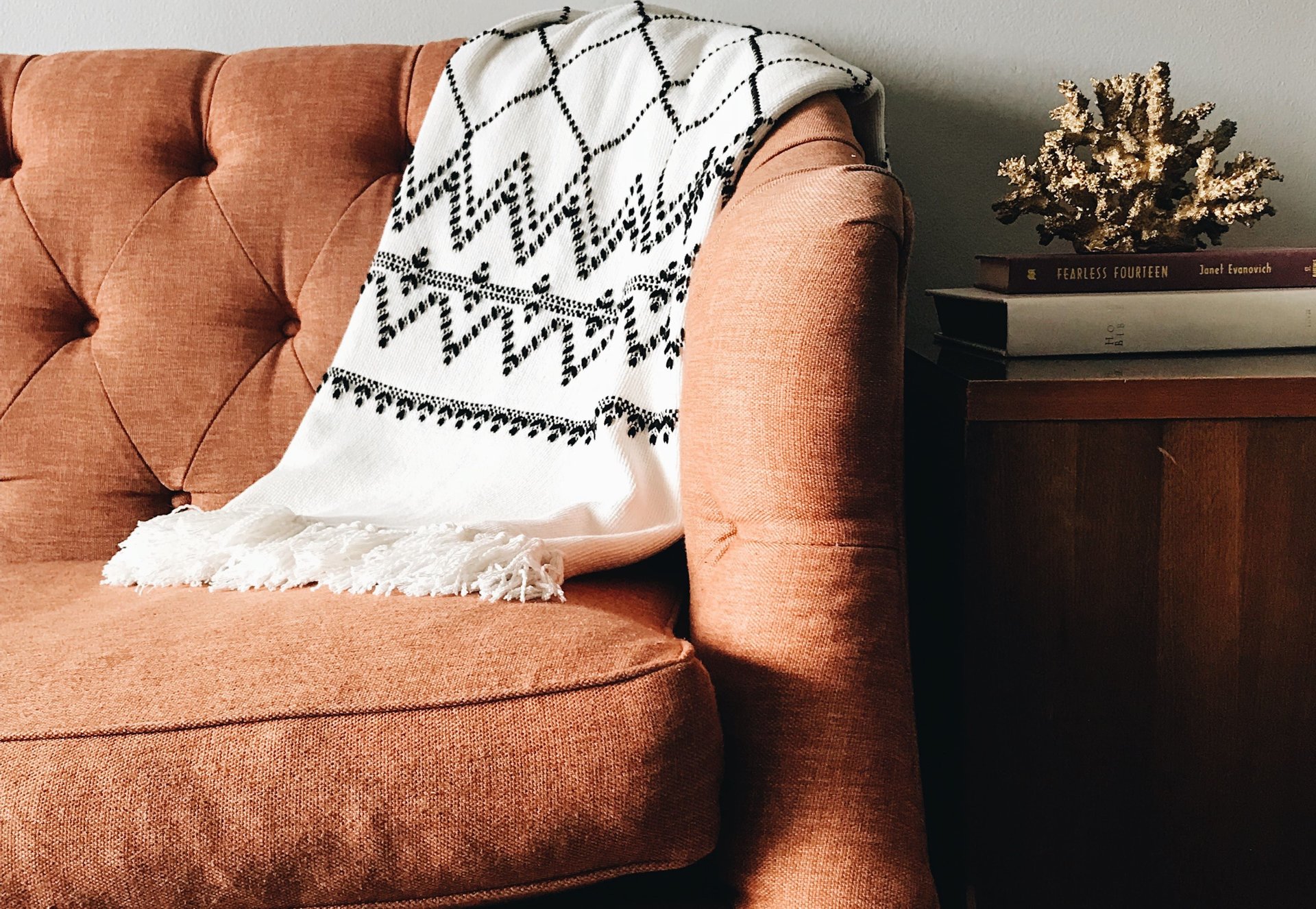 Rust-colored tufted sofa with a patterned white throw beside books and dried flowers.