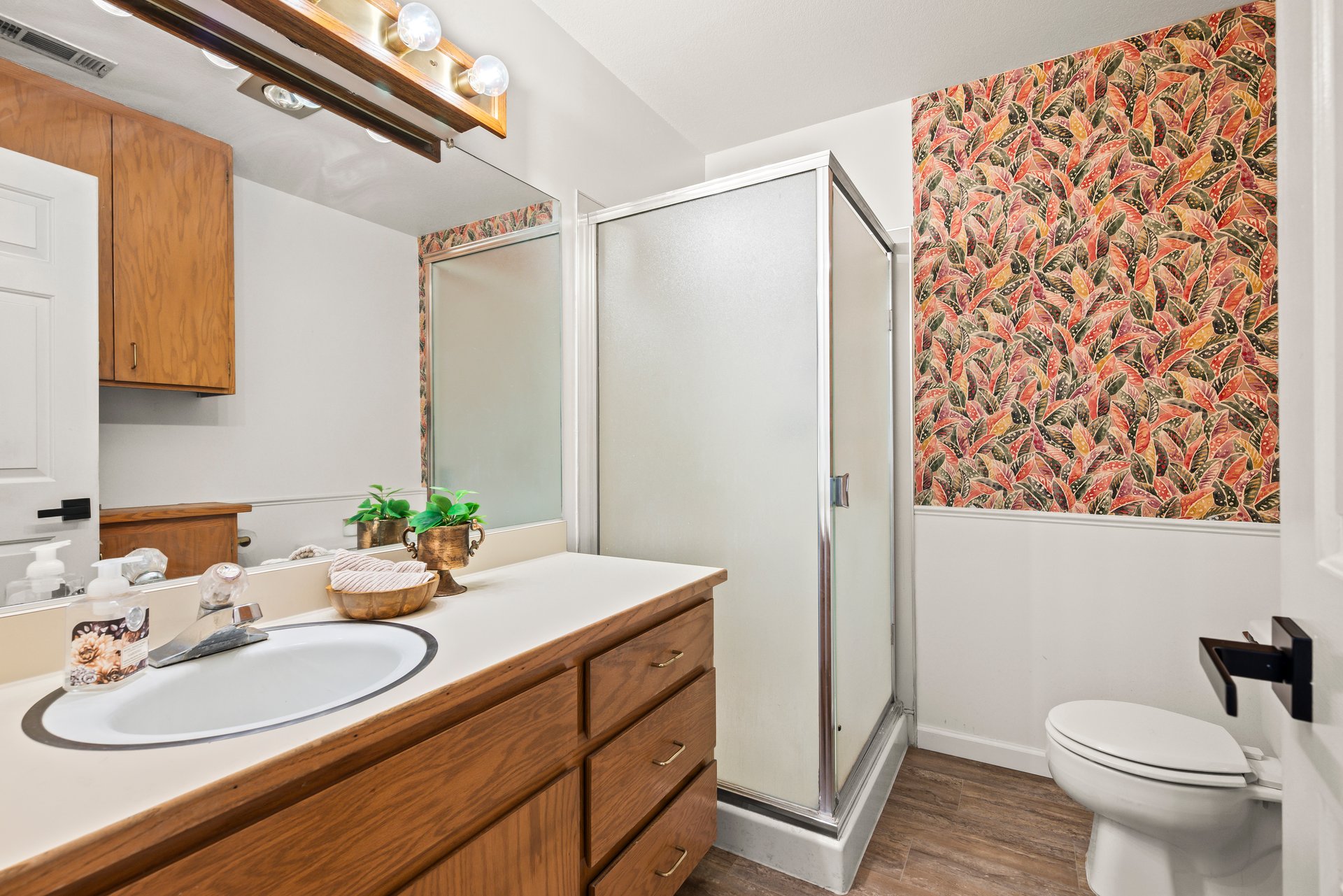 Bathroom with a wood vanity, glass shower, and colorful leaf-patterned wallpaper.