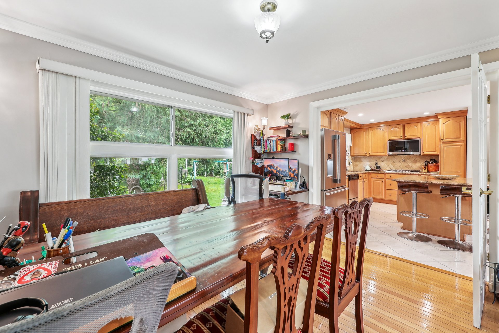 Dining room with a long wooden table, large garden-facing windows, and an adjoining wood-cabinet kitchen.