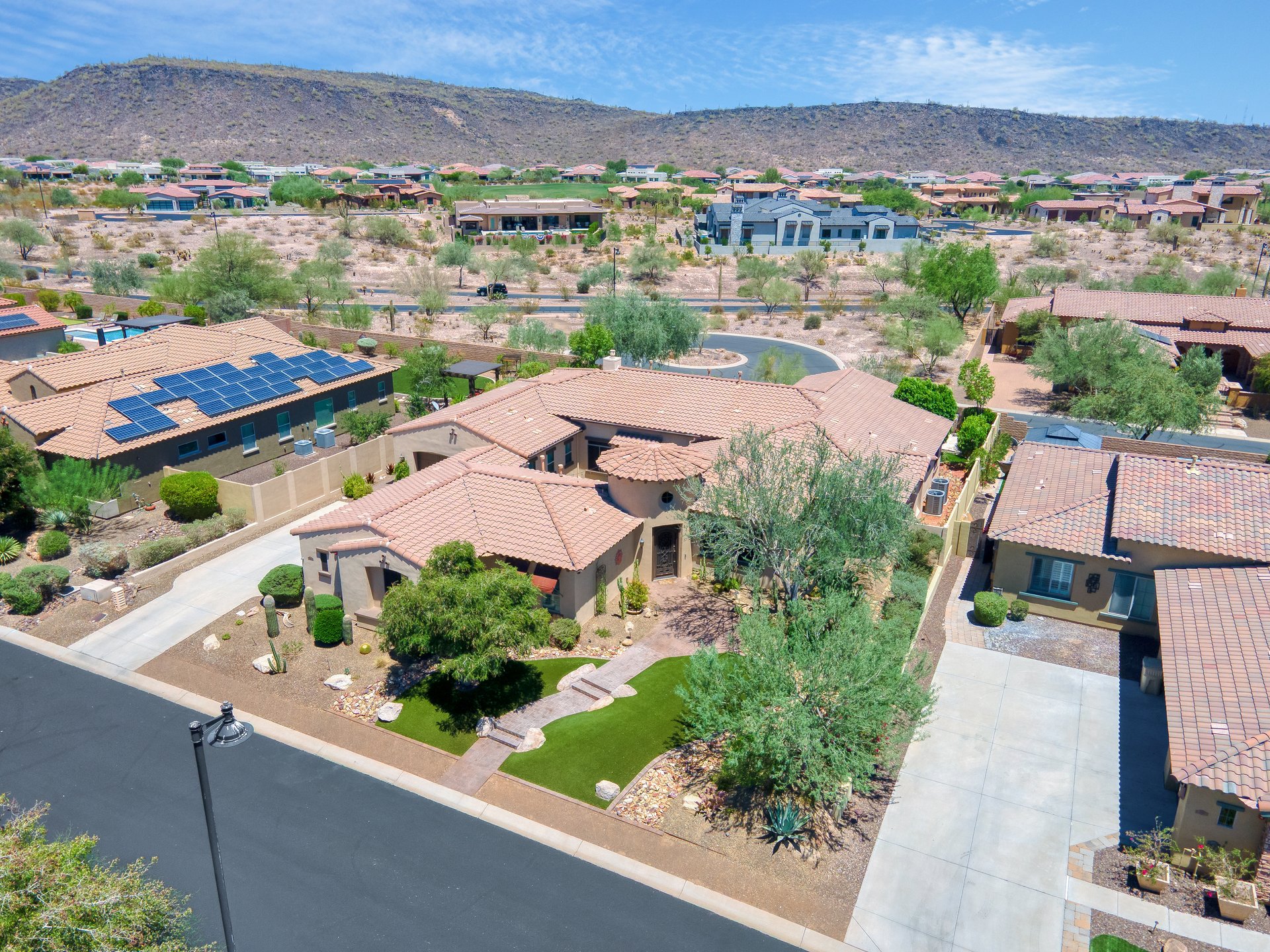 High aerial view of a desert suburban neighborhood with stucco homes, tiled roofs, landscaping, and mountains in the background.