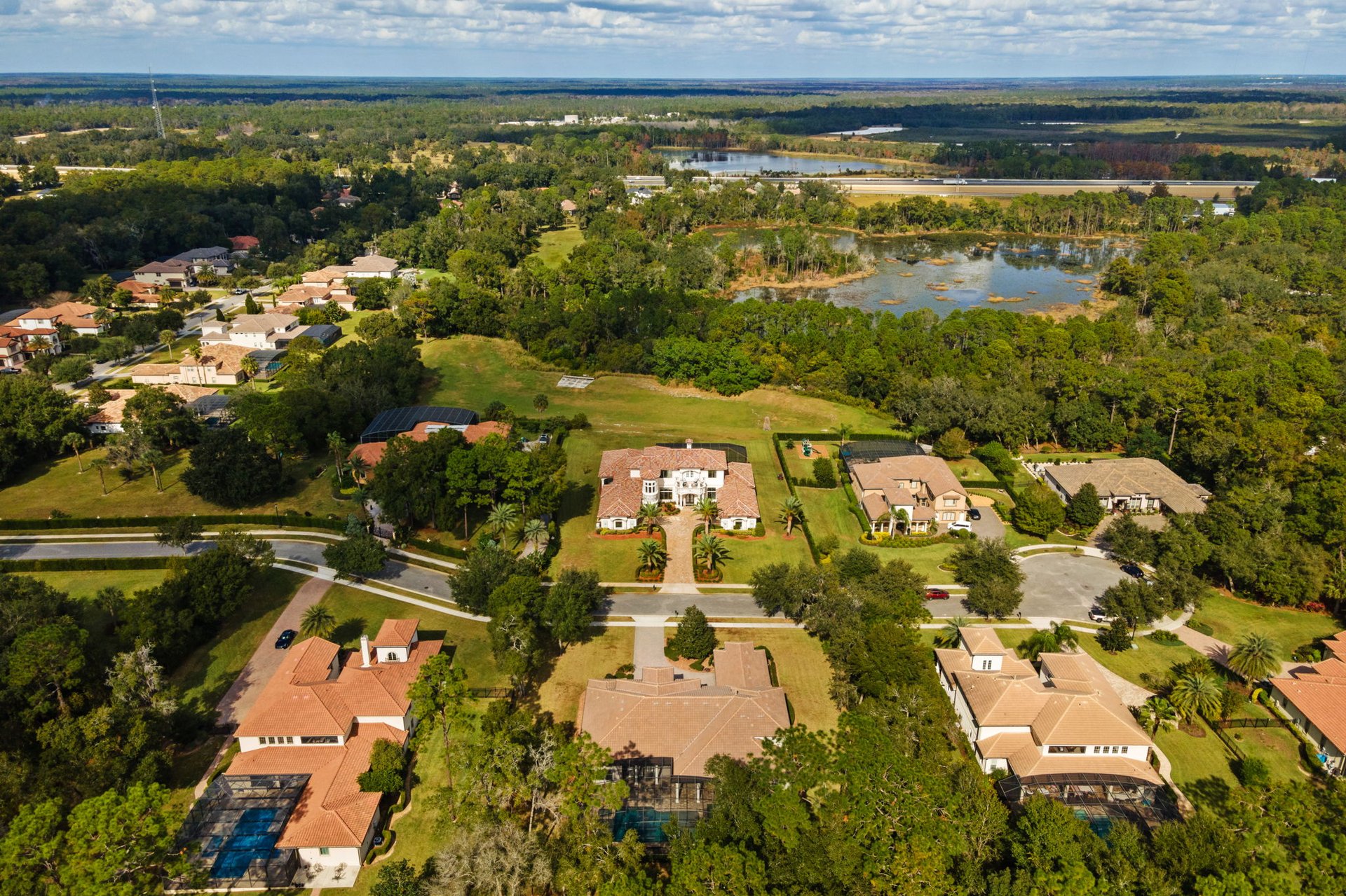 Aerial view of a large tile-roof home among neighboring houses, pools, wooded areas, and a distant lake.