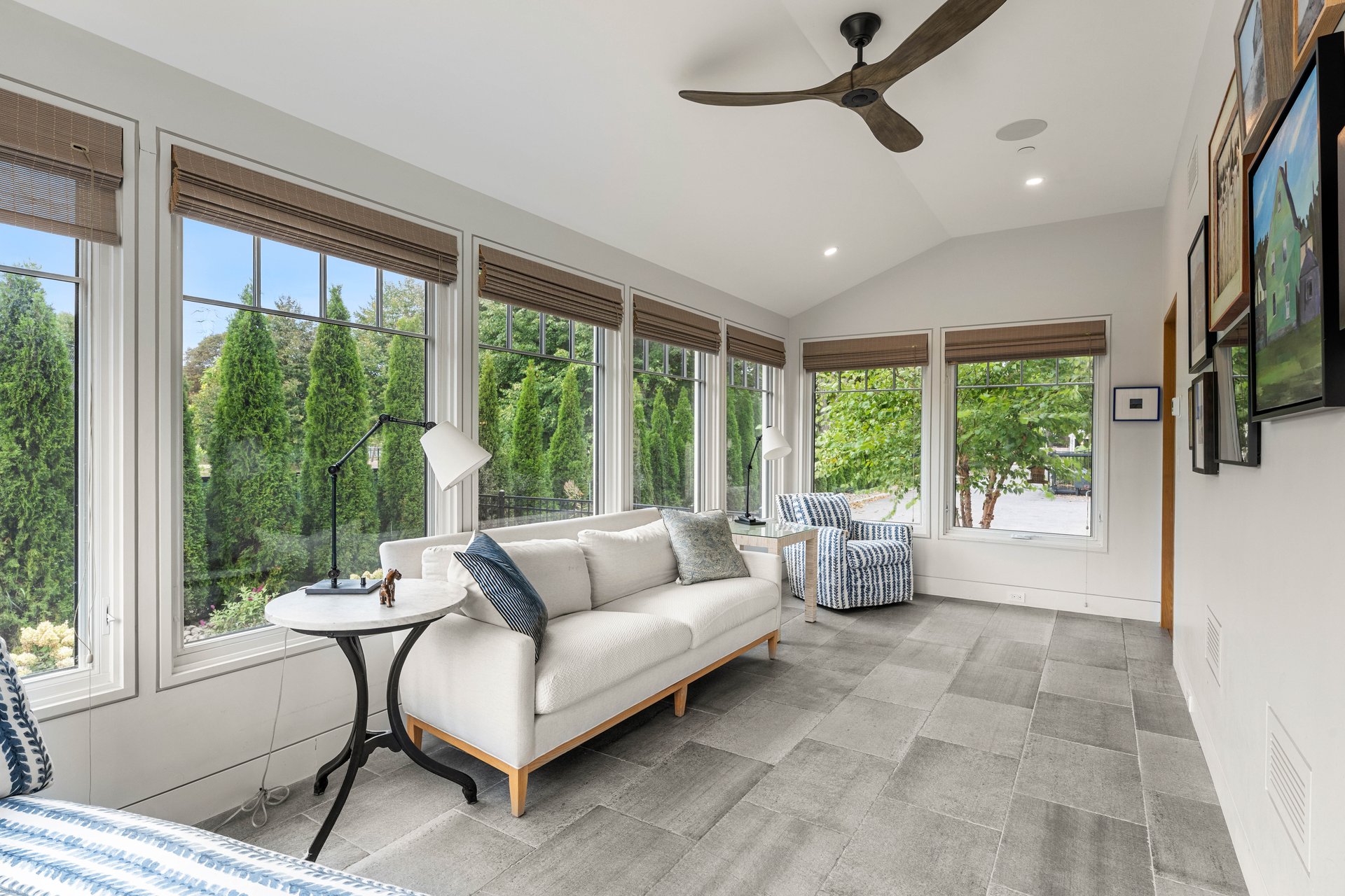 Furnished sunroom with a white sofa, tall garden-facing windows, and gray tile floor.