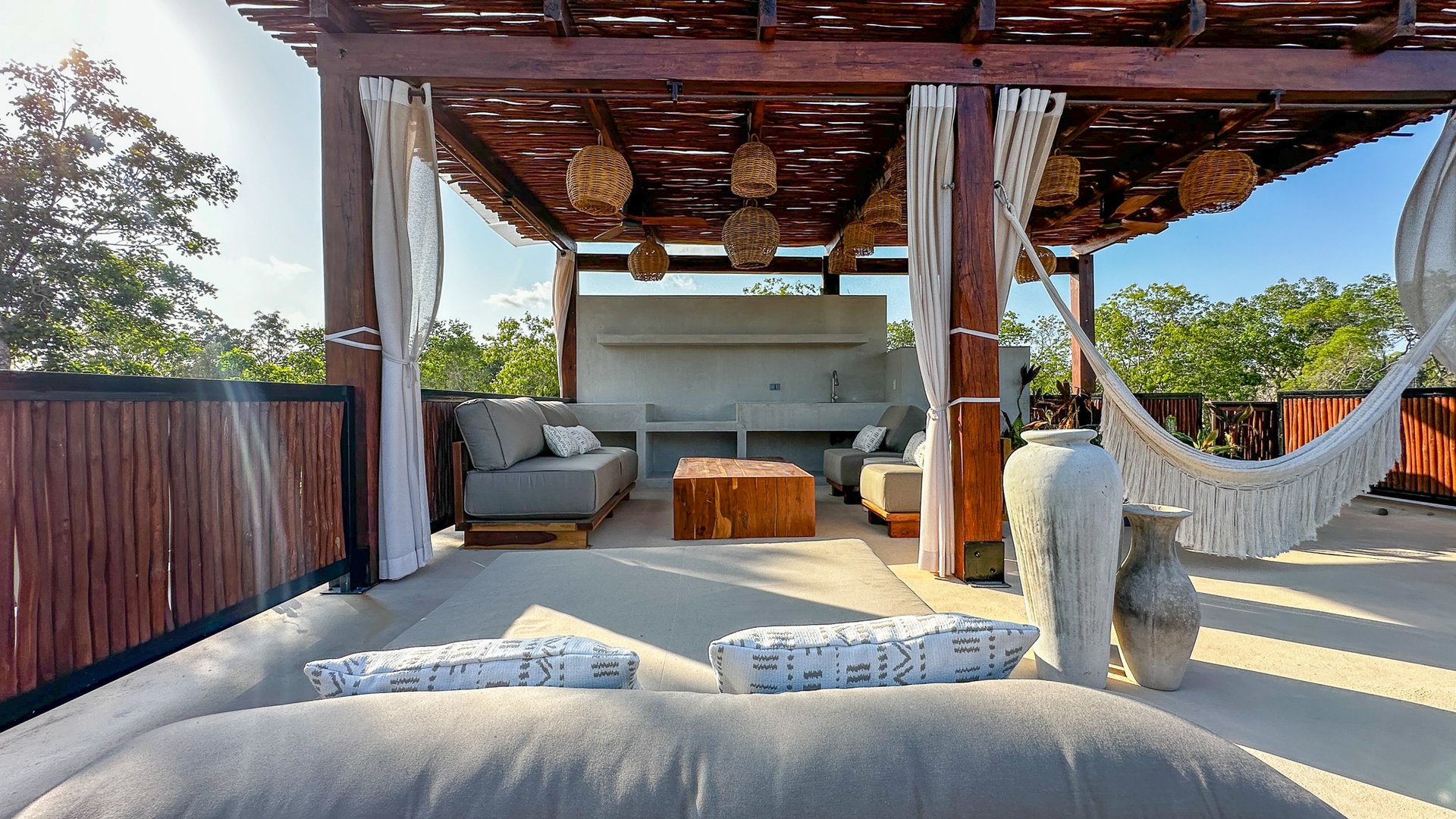 Rooftop lounge with gray sofas, a timber pergola, woven lights, and a white hammock.
