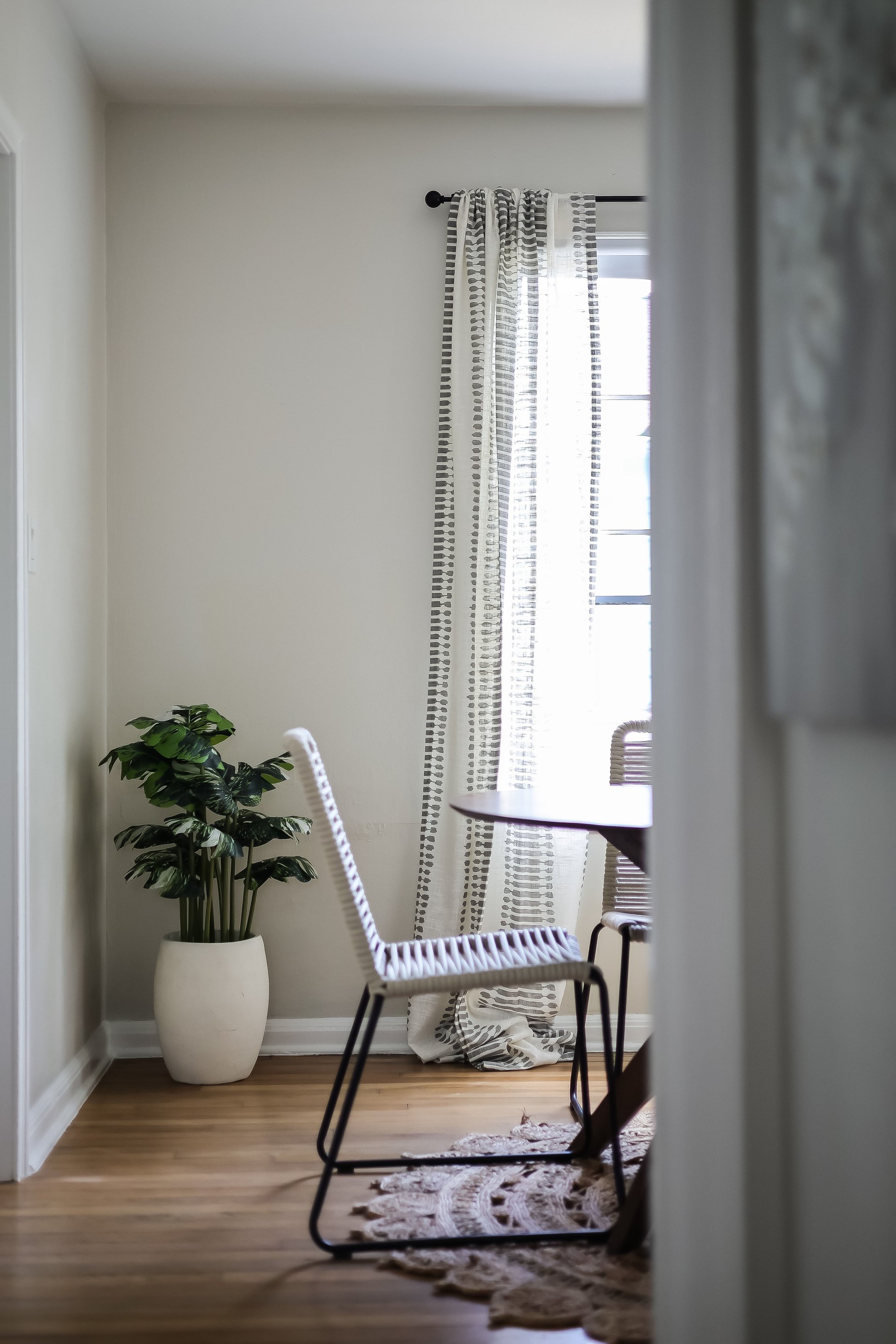 Dining area with woven chairs, patterned curtains, and a potted plant beside a bright window.