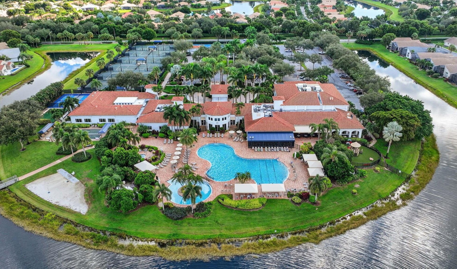 Aerial view of a community clubhouse with pools and tennis courts beside surrounding waterways.