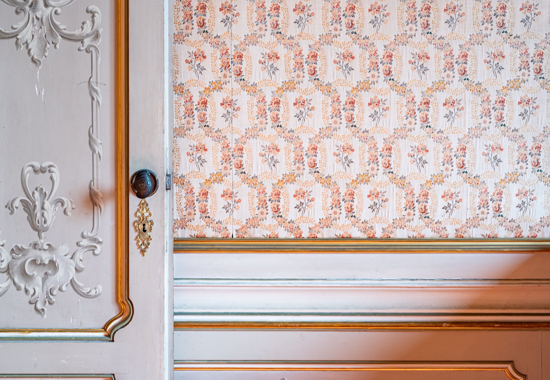 Ornate interior wall with floral wallpaper, sculpted white paneling, gold trim, and decorative door hardware.